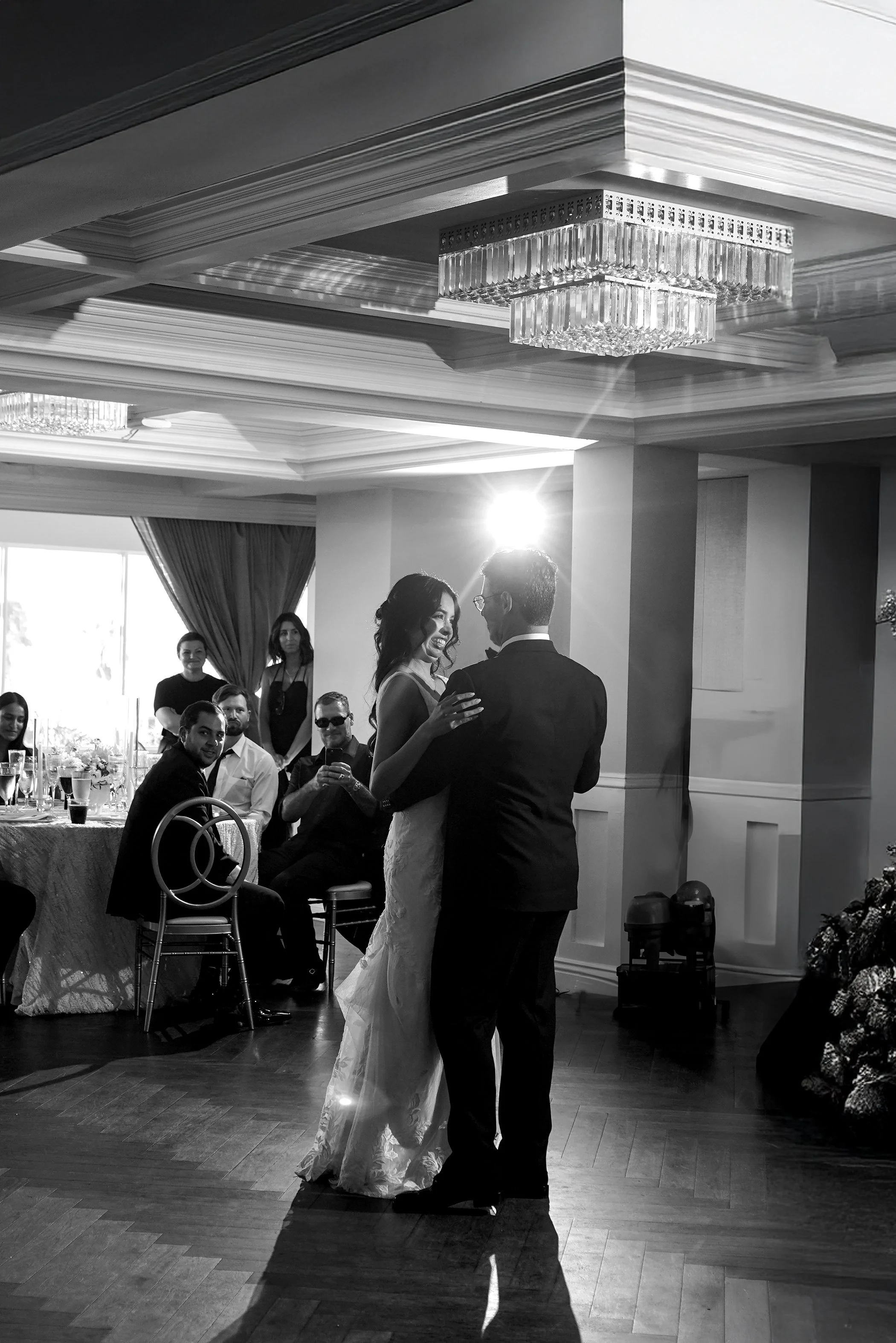 "Newlyweds sharing an intimate first dance in the center of a ballroom, with a bright spotlight creating a lens flare and guests watching from their tables in the background.