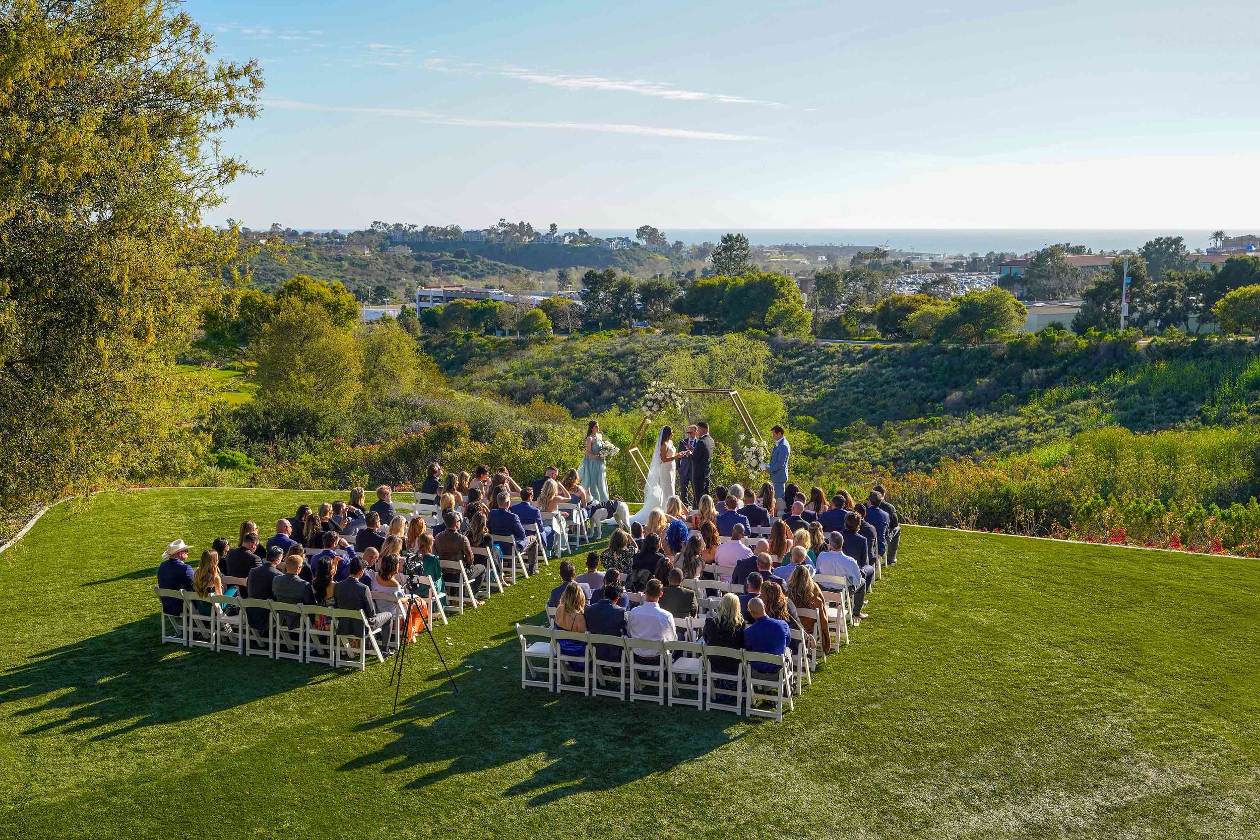 A wide, outdoor photograph of a daytime wedding ceremony on a grassy lawn at The Crossings at Carlsbad in California. Guests are seated on white folding chairs, facing the altar where the bride and groom stand. The scene features a panoramic view of 