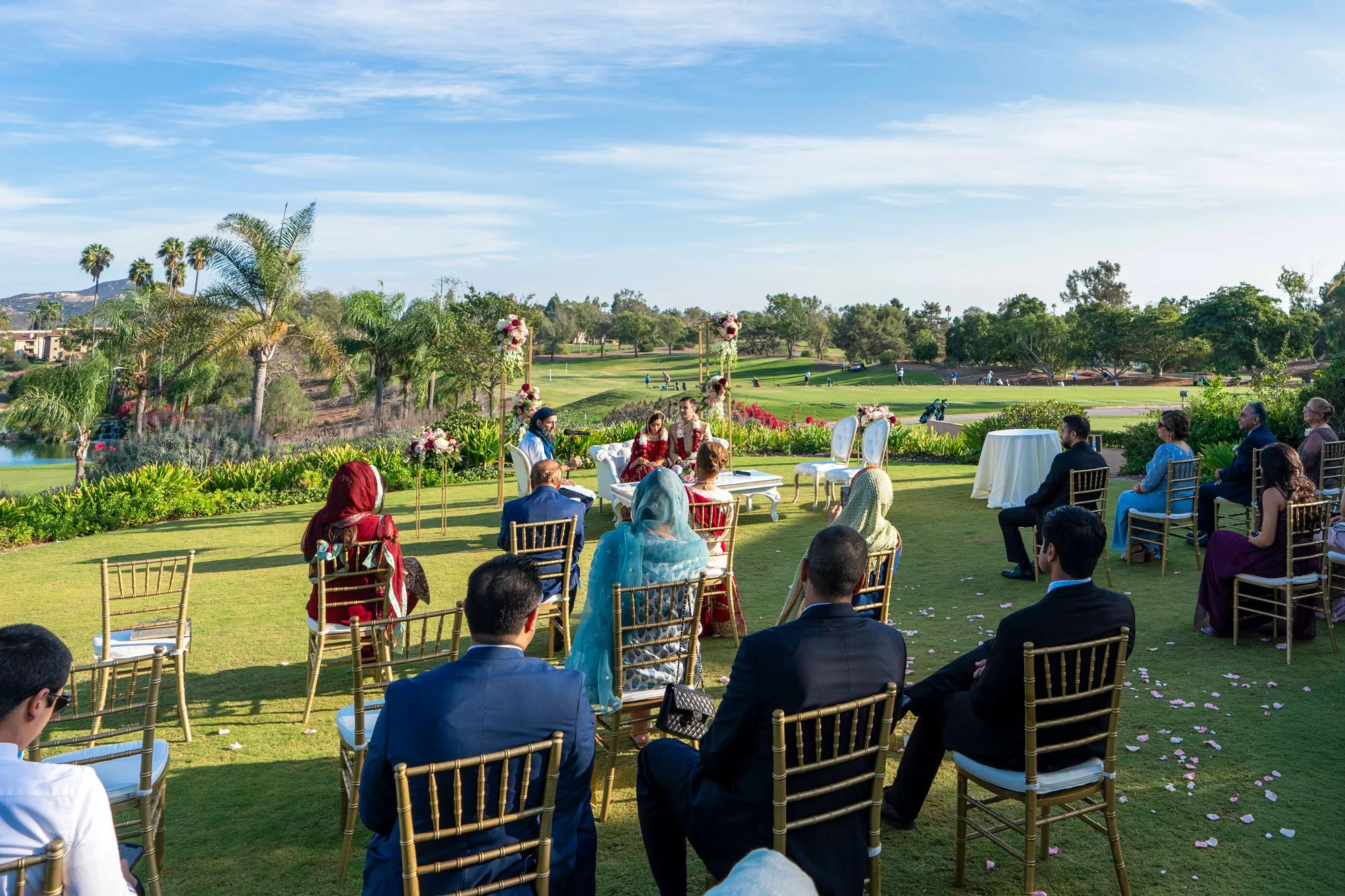 An outdoor photograph of a wedding ceremony taking place on a grassy area, likely a golf course, under a sunny sky. Guests are seated in gold chairs with their backs to the camera, facing the front where the bride and groom are seated at an altar are