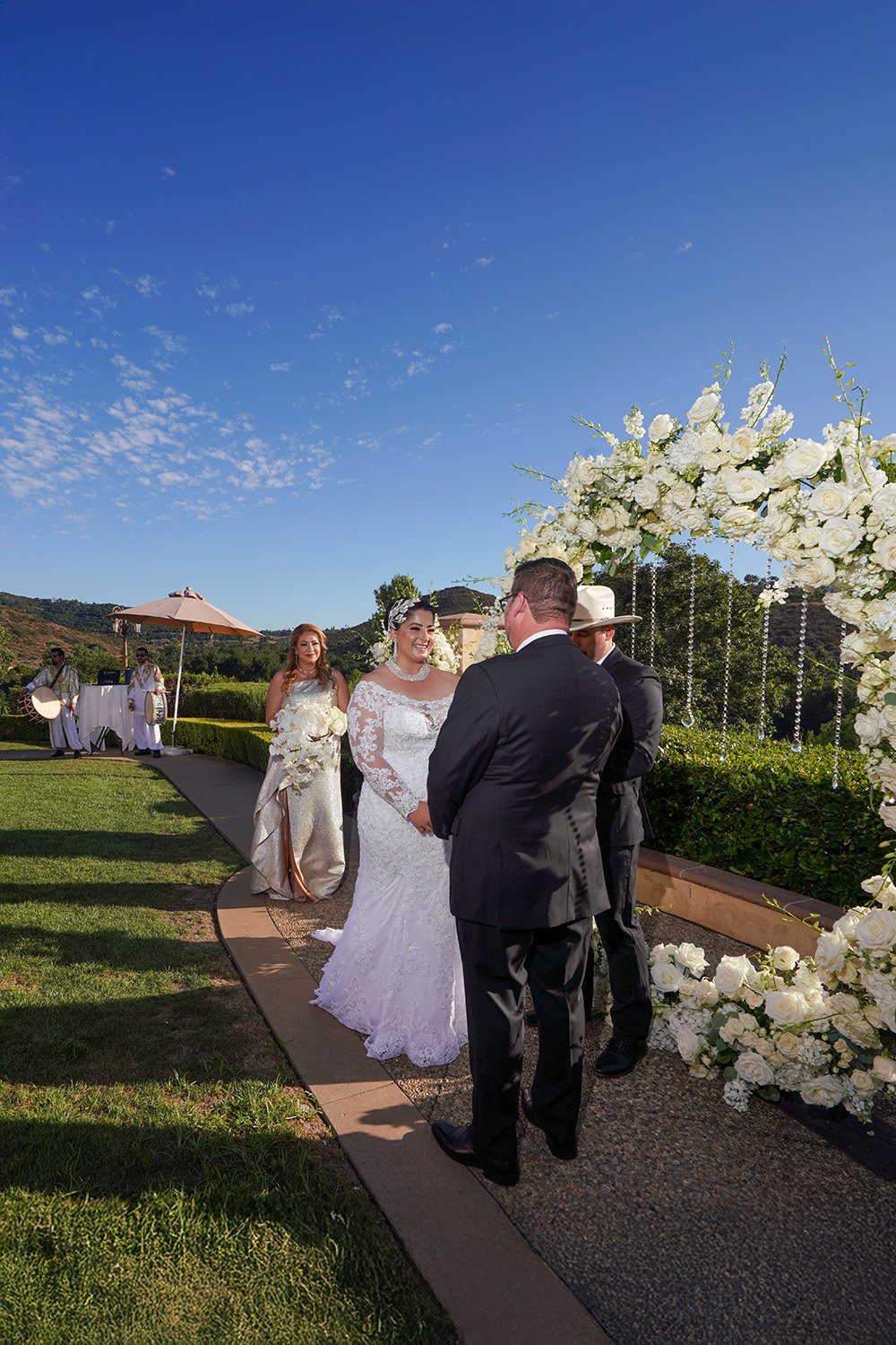 A bride and groom stand at the altar during an outdoor wedding ceremony. The bride, in a long-sleeved lace gown and jeweled headpiece, faces the groom, who is in a black tuxedo and seen from behind. An officiant in a cowboy hat stands beside them und