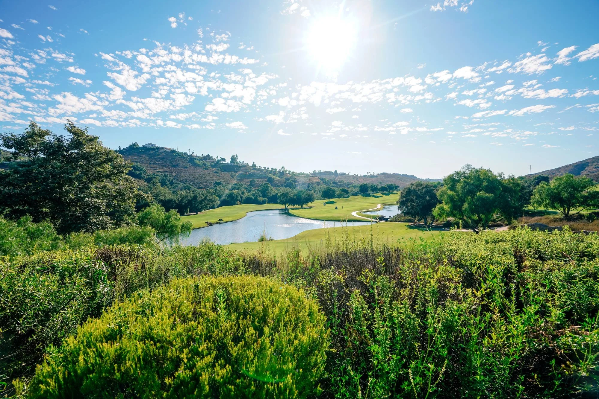 A wide, outdoor photograph of a lush green Maderas golf course landscape under a bright sunny sky, featuring a large pond and rolling, tree-covered hills in the background.



