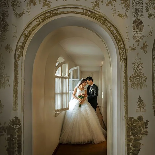 A bride and groom share an intimate moment in an elegant, arched hallway. The bride, in a voluminous white ballgown, holds a colorful bouquet while the groom in a dark suit leans in close. 