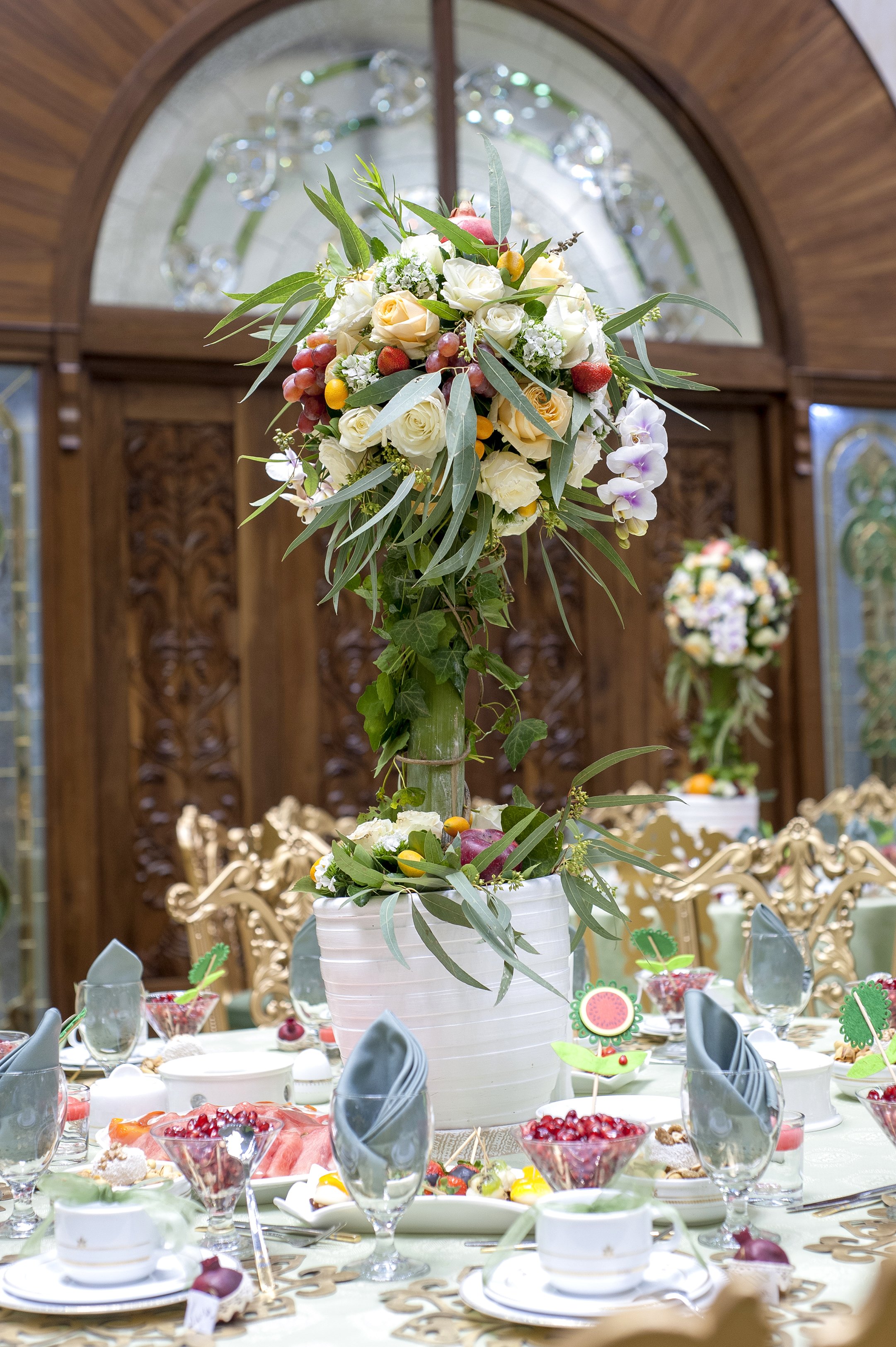 A formal dining table setting for an event, featuring a tall central floral and fruit arrangement with grapes, roses, and greenery, place settings with folded blue napkins, bowls of pomegranates and fruit, and a carved wooden door backdrop.
