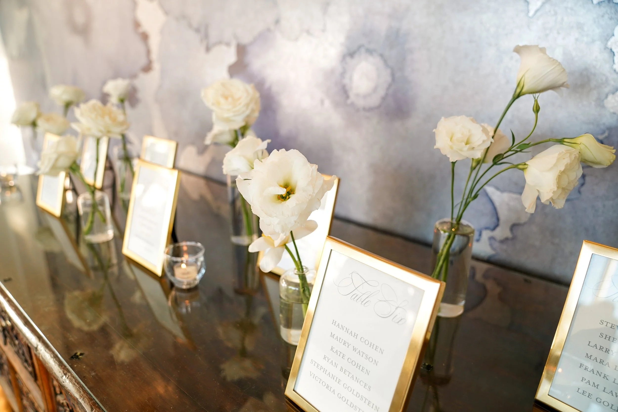 A dark wooden credenza displaying several gold-framed event table assignment cards with guest names, accompanied by small glass vases of white flowers and votive candles.