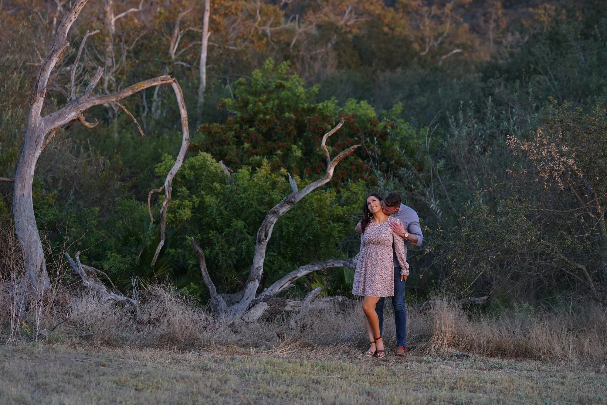 An outdoor photograph of a couple embracing in a natural, overgrown area during warm evening light. The woman wears a short, light-colored, floral dress, and the man, in a grey shirt and blue jeans, stands behind her, appearing to kiss her on the nec