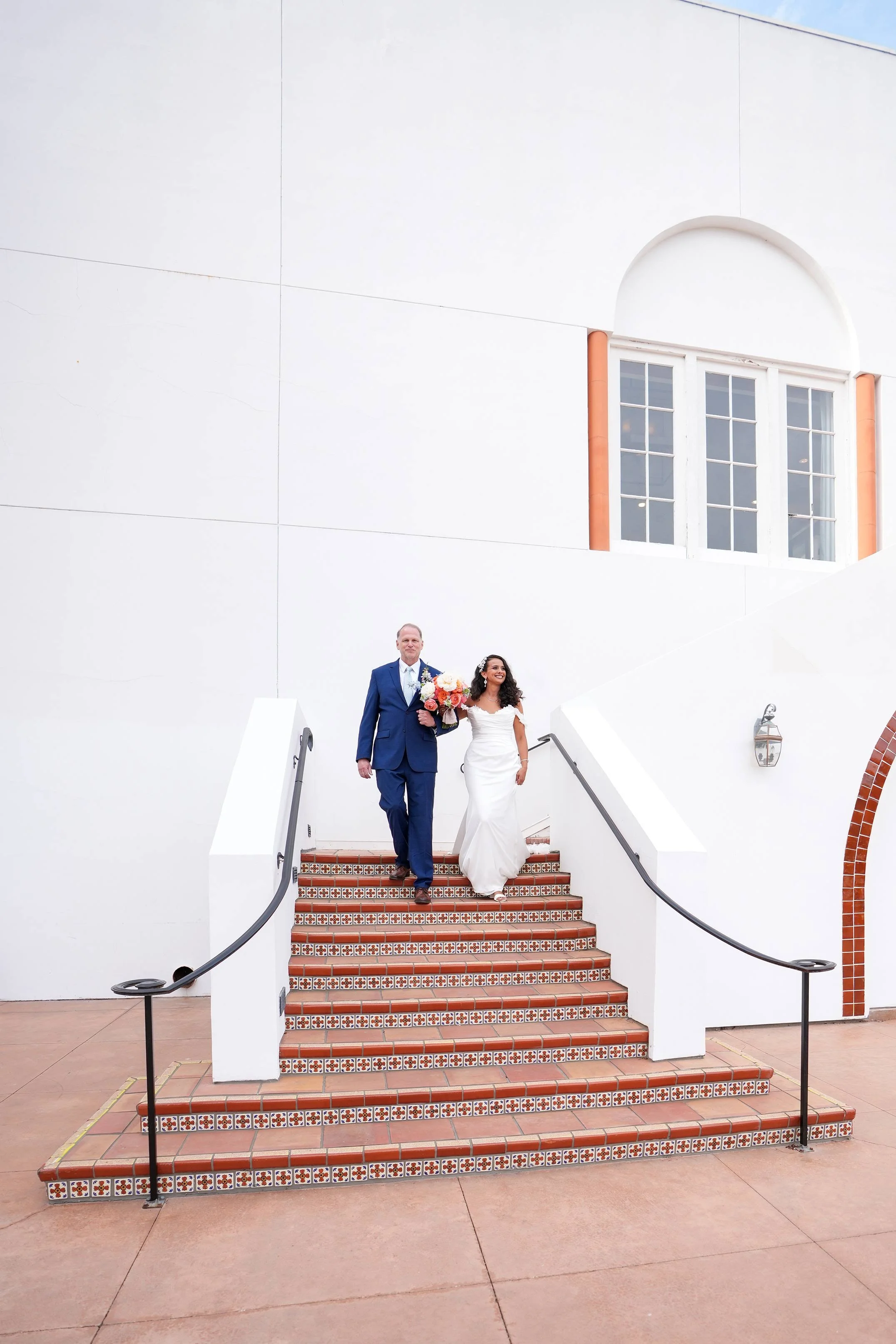 A bride in a white wedding gown holding a bouquet and her father in a blue suit walk down a set of outdoor stairs adorned with patterned red and brown tiles in Omni La Costa, beside a white stucco building.