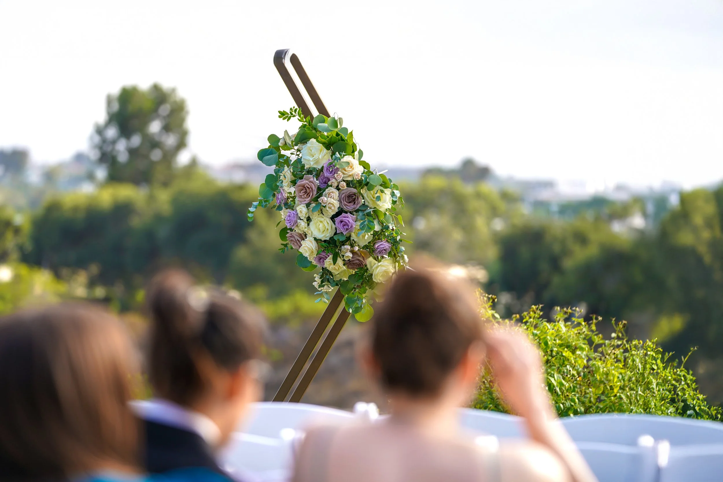A rustic yet elegant floral arrangement of white, cream, and lavender roses with eucalyptus greenery, attached to a dark metal frame, with a blurred background of an outdoor ceremony setting and guests.
