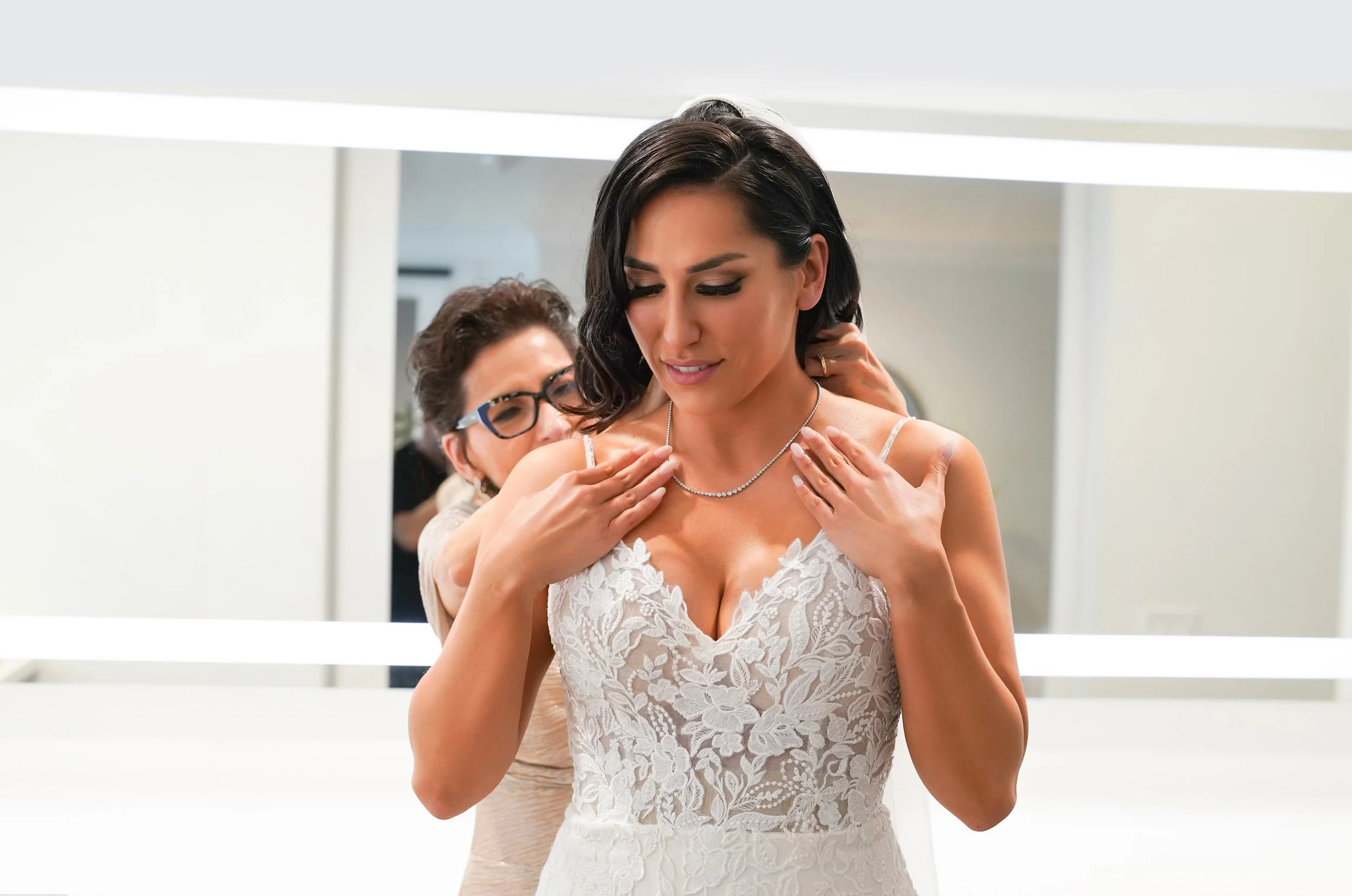 An indoor photograph of a bride in a white lace wedding dress with a deep V-neckline, getting help with a diamond or crystal necklace. An older woman wearing glasses stands behind her, assisting with the clasp in a modern room with white walls and mi