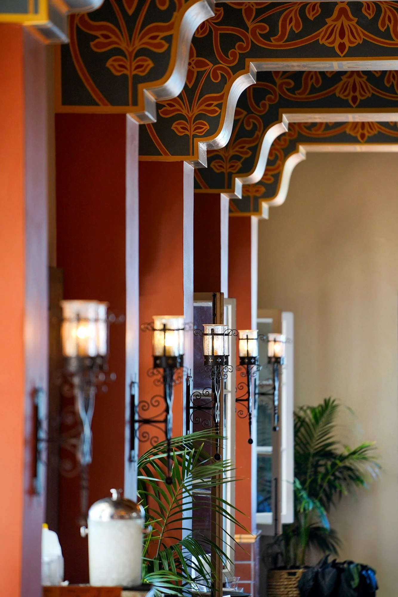An indoor photograph of an elegantly decorated hallway with rust-colored walls and an ornate, painted ceiling featuring dark blue, red, and gold designs with wavy silver trim. Several lit wrought iron wall sconces are mounted on the walls, and green 