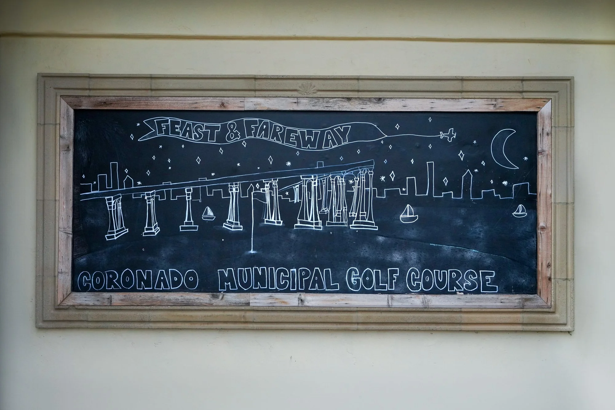 A chalkboard sign featuring a white line drawing of a stylized skyline with the San Diego-Coronado Bridge, boats, a moon, and an airplane, alongside the text "FEAST & FAREWAY" and "CORONADO MUNICIPAL GOLF COURSE".