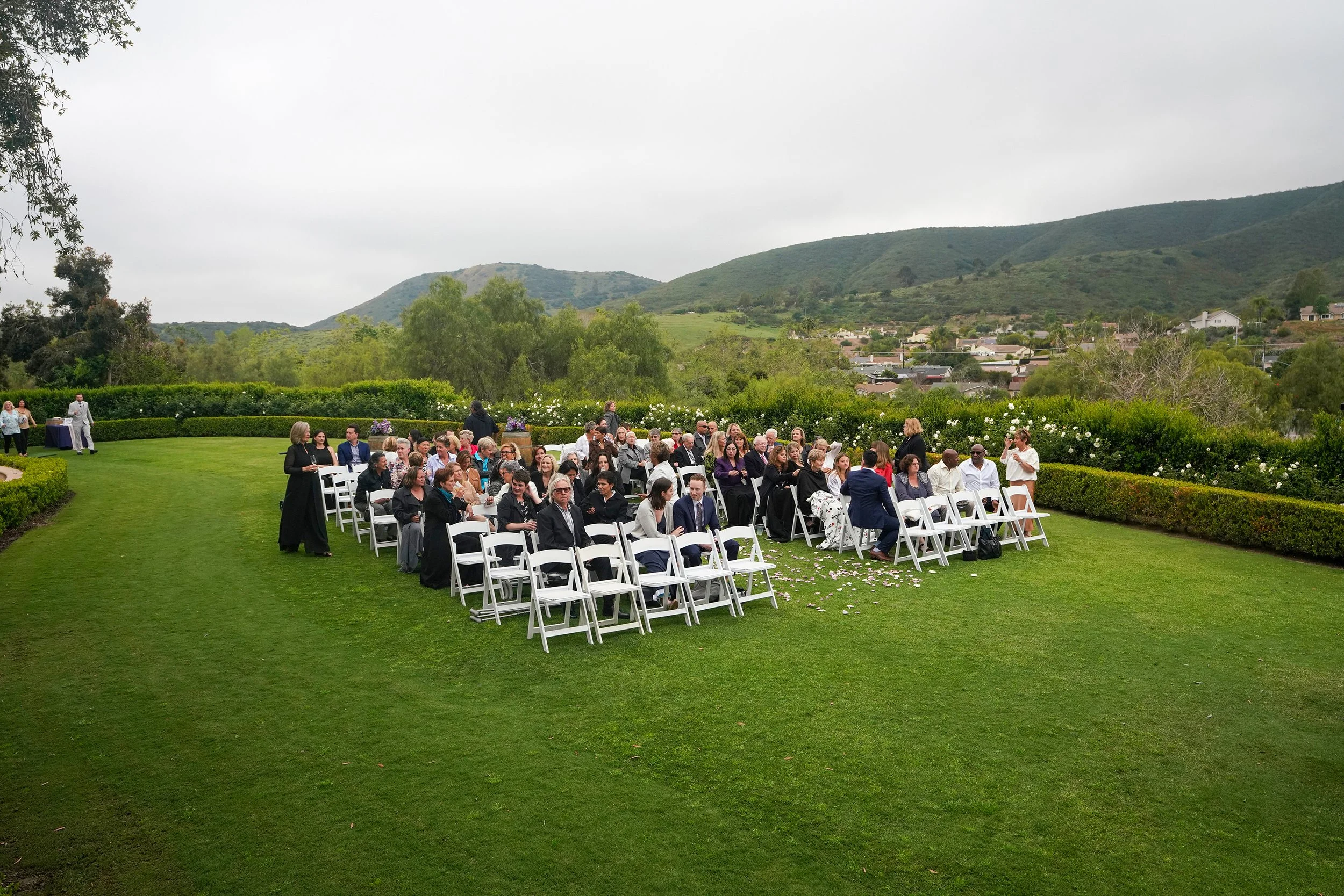 An outdoor wedding ceremony taking place on a green lawn, with guests seated in white chairs, surrounded by manicured hedges and rolling hills in the background under an overcast sky.