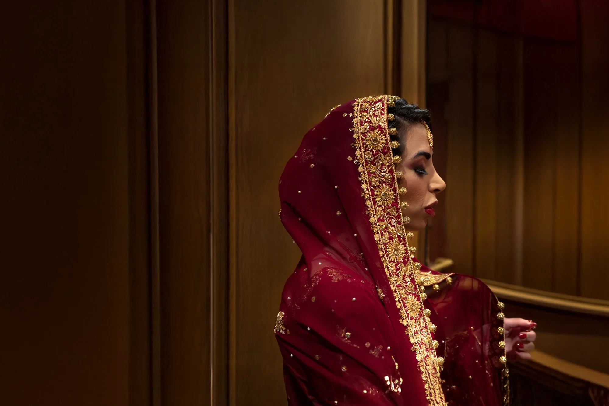 A side profile photograph of a bride wearing a deep red, gold-embroidered traditional Indian wedding outfit and jewelry, standing indoors against wood paneling.


