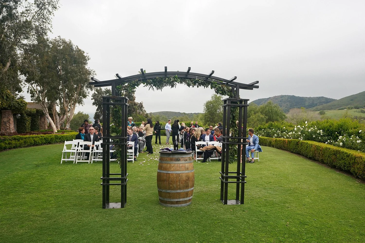 An outdoor wedding ceremony setup on green grass, featuring a black metal arch decorated with white and pink roses and greenery, and a wooden wine barrel serving as a small table.