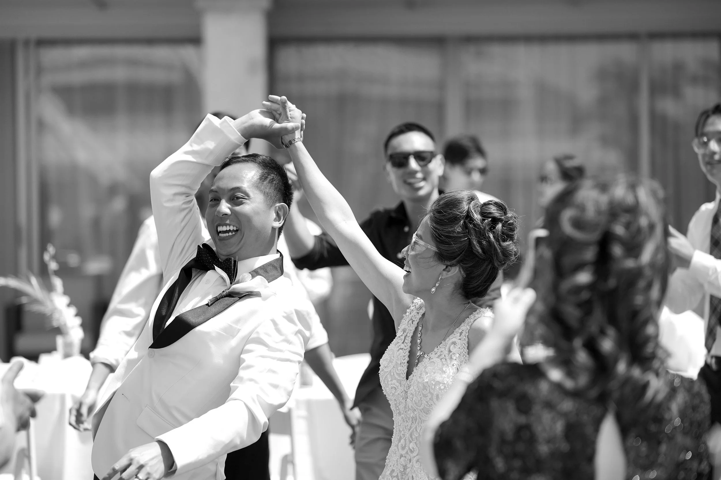 A black and white photograph captures a vibrant moment of a bride and groom laughing and dancing with their hands raised in the air during their outdoor wedding reception at a Mission Bay resort.