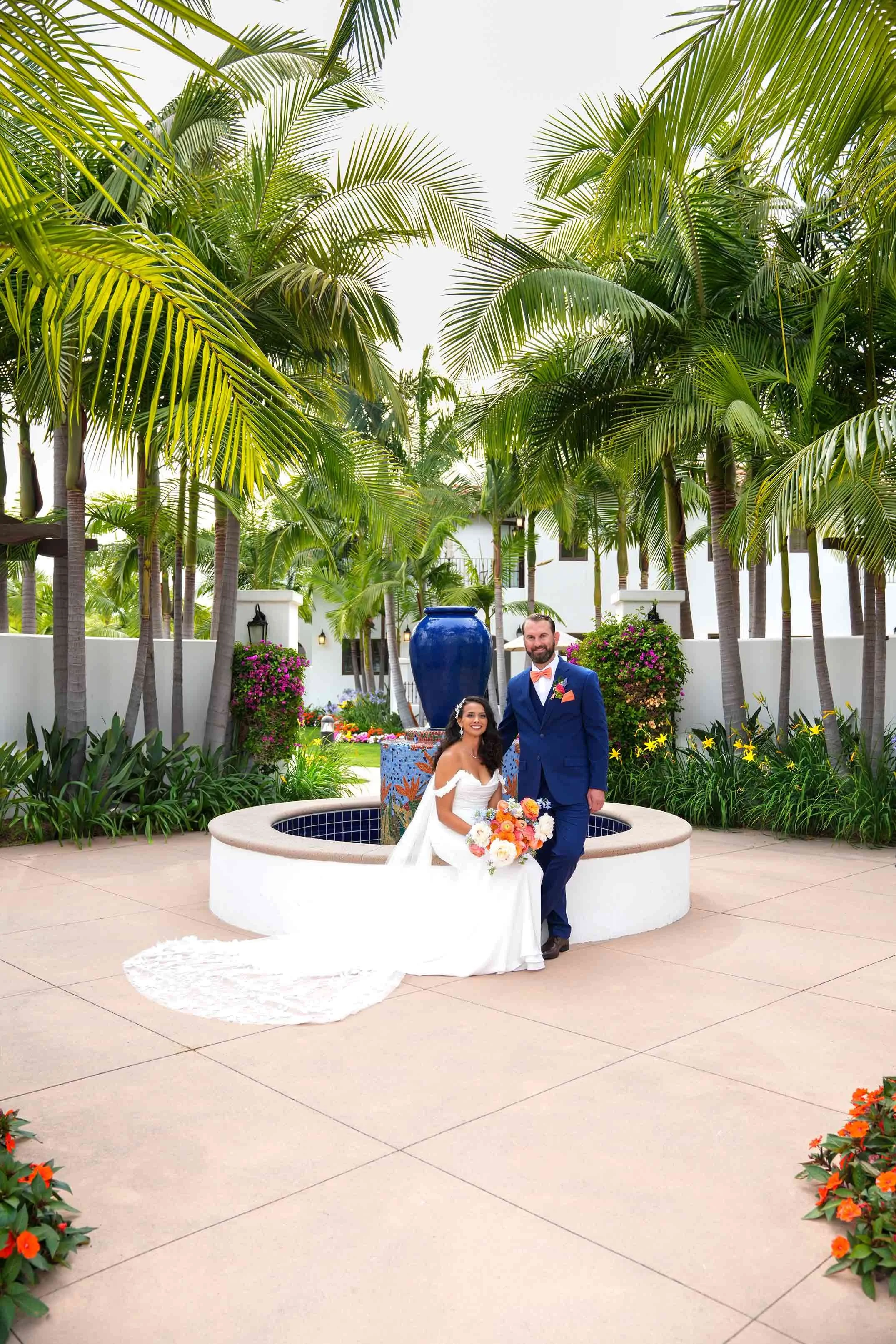 A bride in a white, off-the-shoulder gown and a groom in a blue suit sit by a circular white fountain surrounded by lush palm trees at the Omni La Costa Resort & Spa.