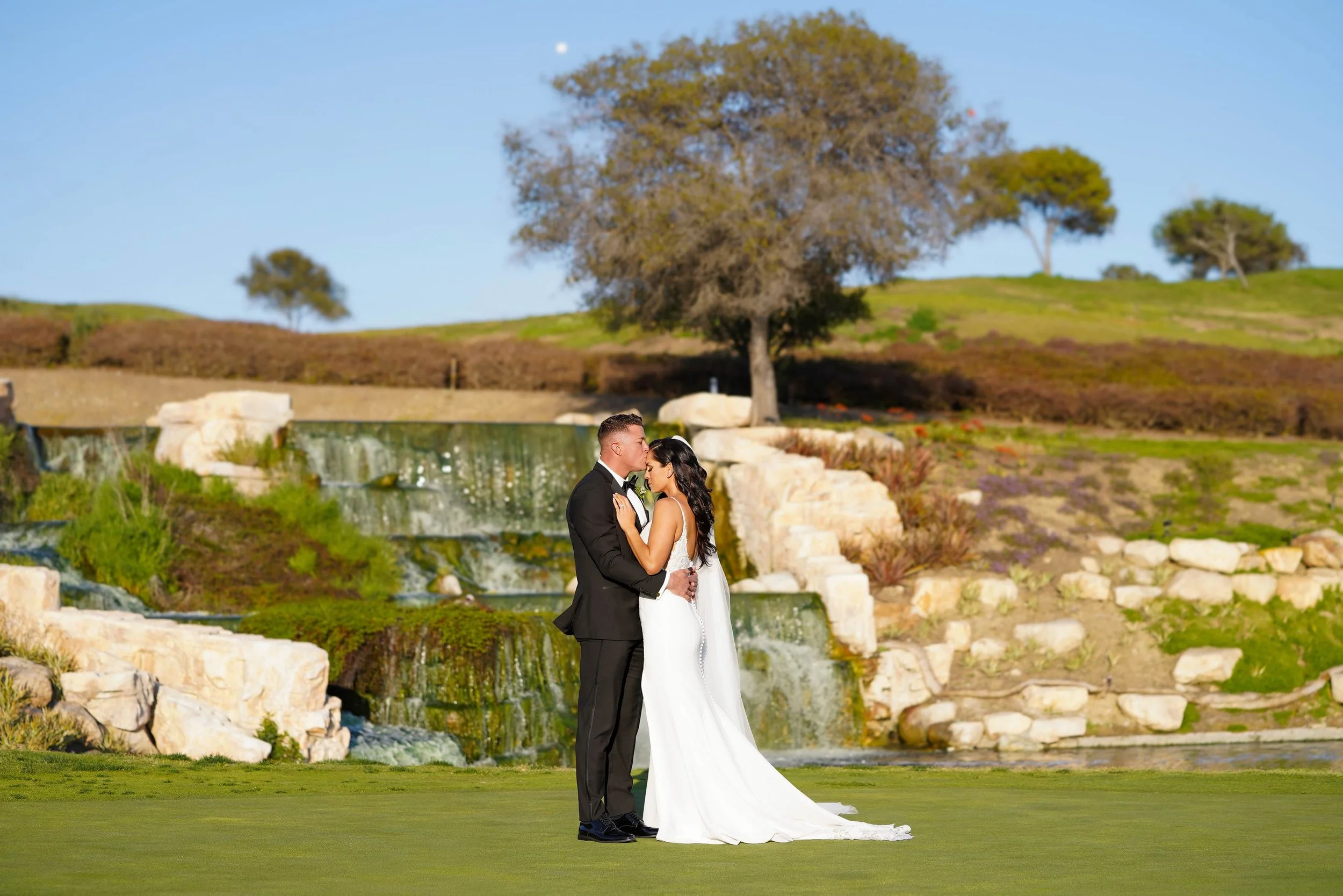 An outdoor photograph of a bride and groom embracing and kissing on a grassy lawn. The groom wears a black tuxedo and bow tie, and the bride wears a white wedding dress with a low back and long train. They are standing in front of a man-made rock wat