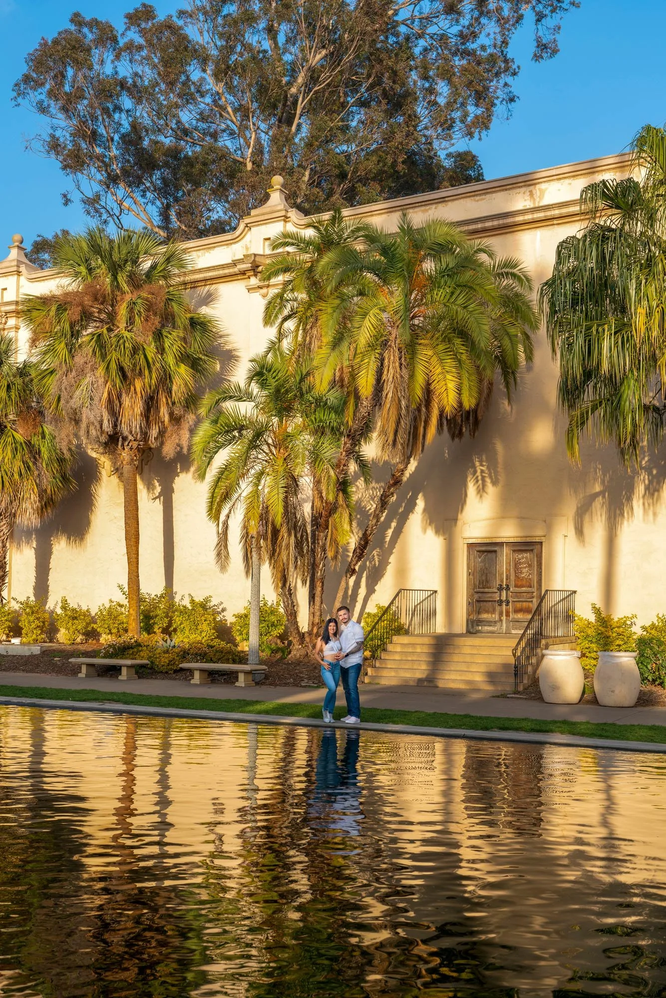 A couple standing by a body of water with a large beige building with tropical trees in Balboa Park, San Diego, and a clear blue sky in the background.