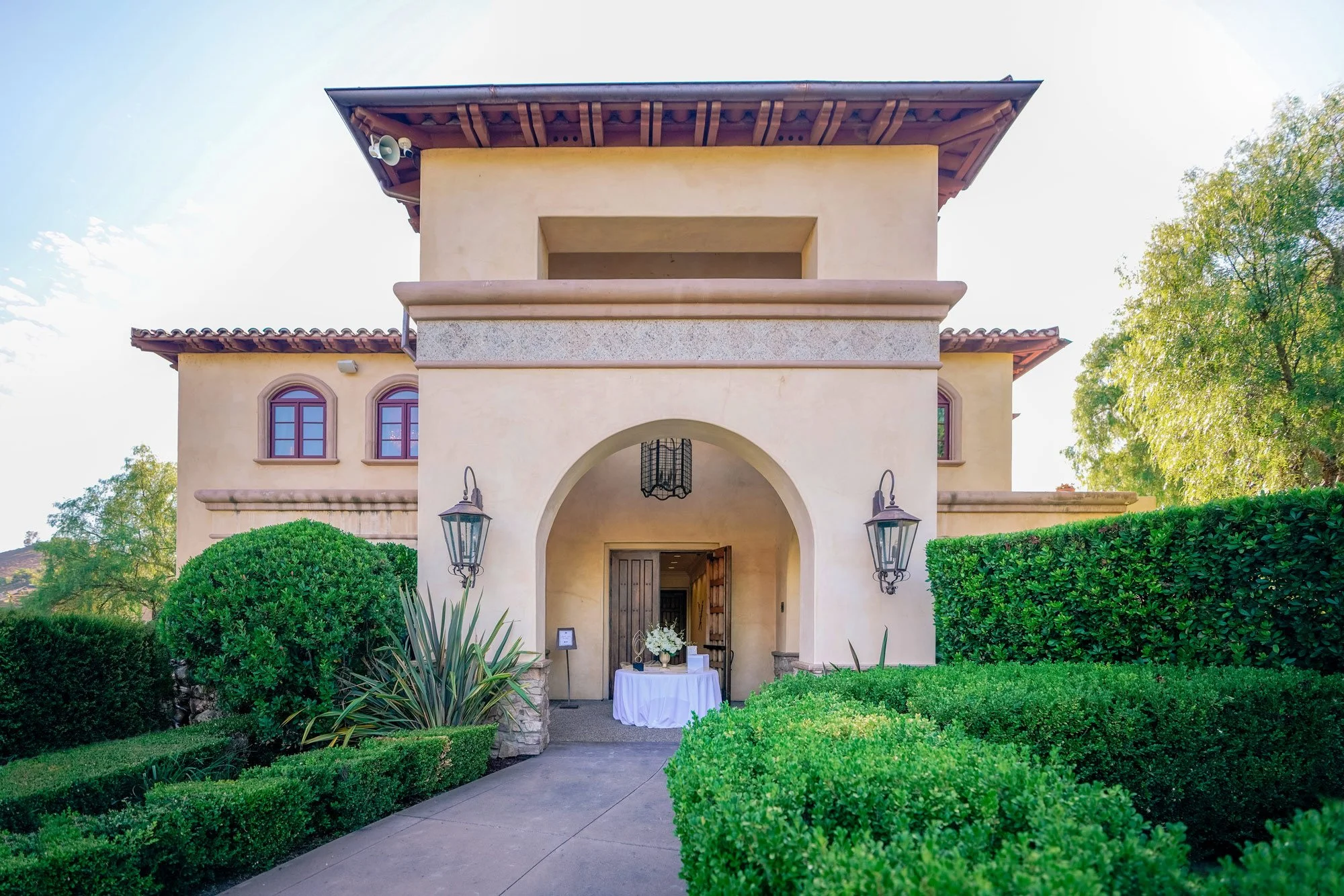 An outdoor photograph of the entrance to a large, two-story building with Spanish-style architecture. The building has a stucco facade, a red-tiled roof, and an arched entryway. The entrance is flanked by two large, wrought-iron lanterns, and lush, n