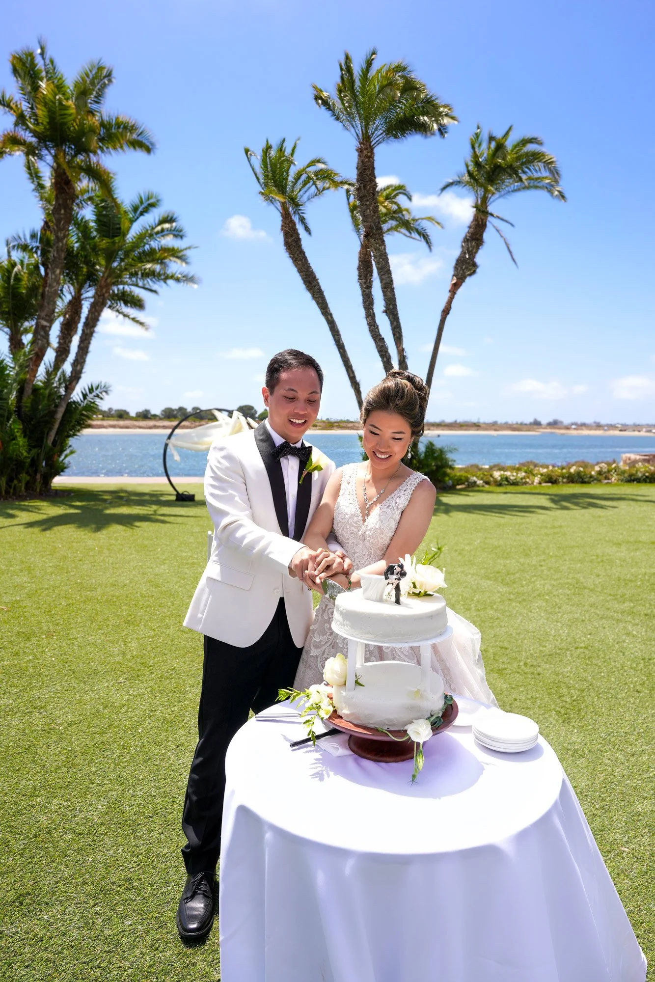 A smiling bride in a white lace gown and a groom in a white tuxedo jacket hold a knife together, cutting into their two-tiered wedding cake during their sunny outdoor reception on a green lawn overlooking the water and framed by tall palm trees in Sa