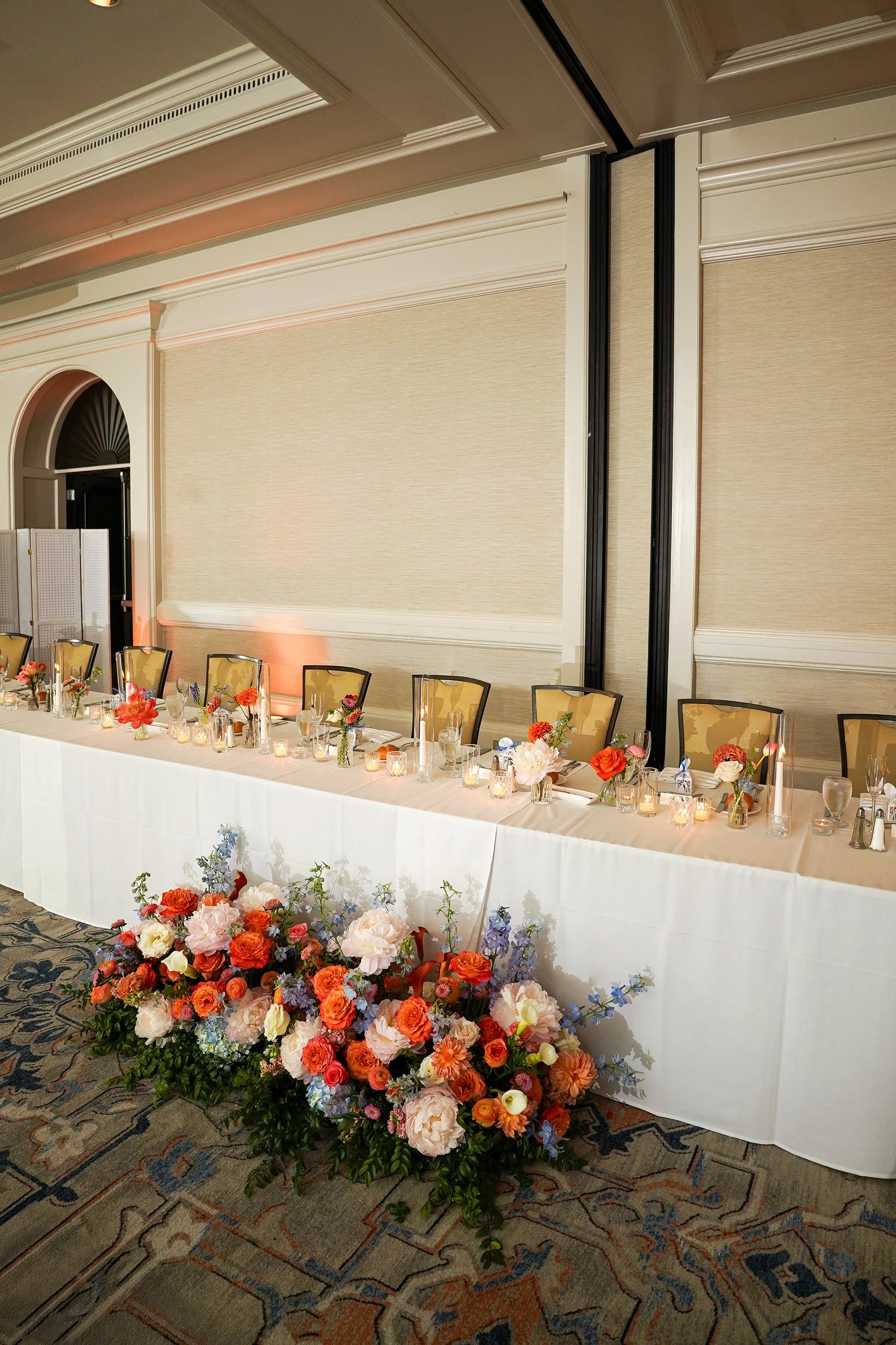 A long, white-clothed wedding head table is elegantly decorated with a vibrant floral runner and multiple small, lighted candles. In the foreground, a large, lush floral arrangement sits on the patterned blue and tan carpet at the foot of the table, 