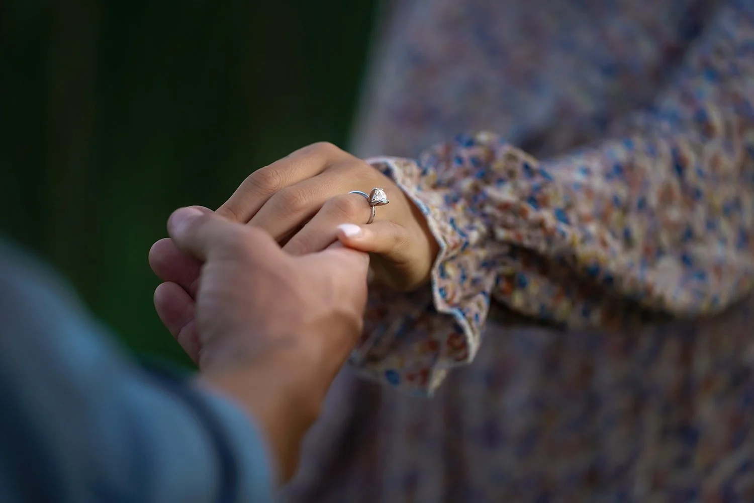 A close-up photograph of a woman's hand, adorned with a diamond engagement ring, being held gently by a man's hand. The woman is wearing a long-sleeved floral-patterned top, and the background is blurred green foliage.