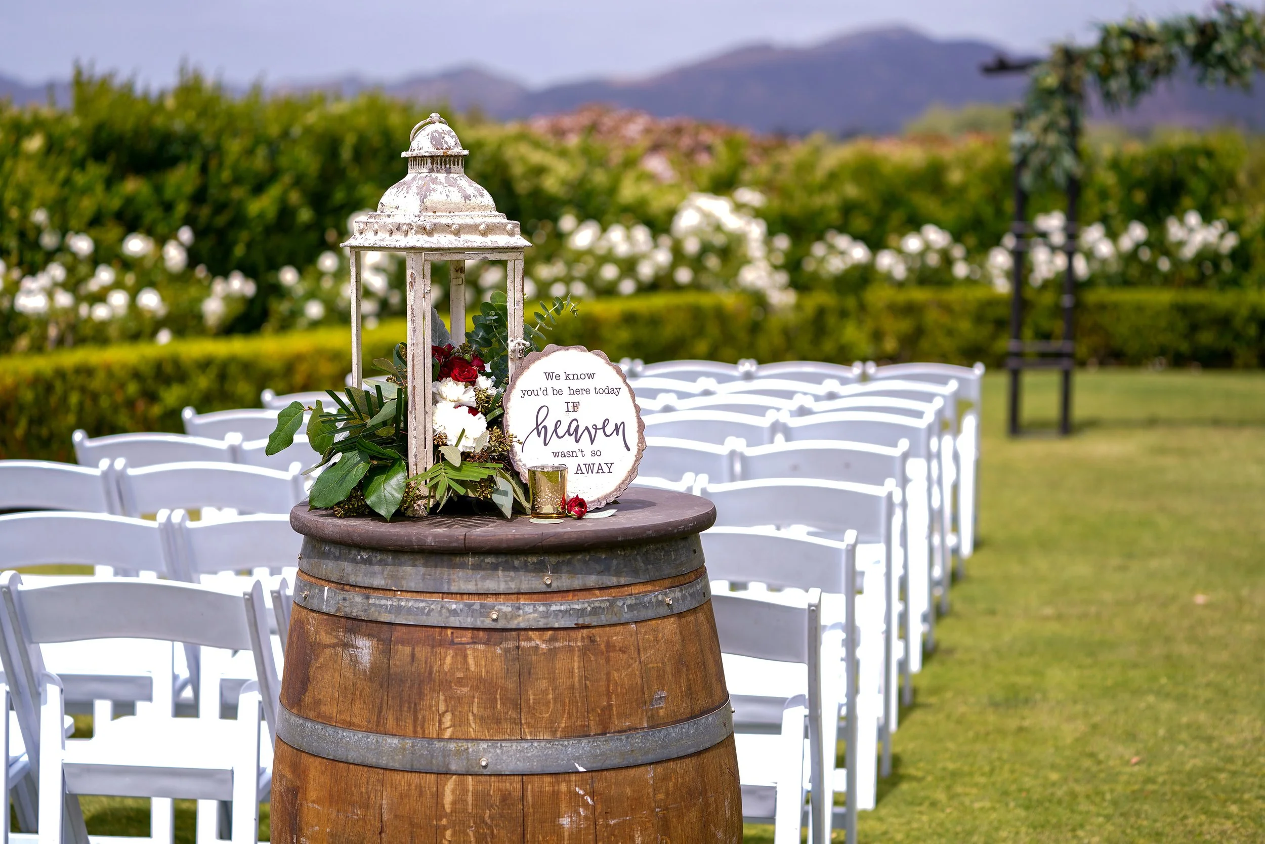 A rustic outdoor wedding ceremony setup featuring a wine barrel as an aisle marker, topped with a white lantern and a memorial sign, with rows of white chairs set on green grass.
