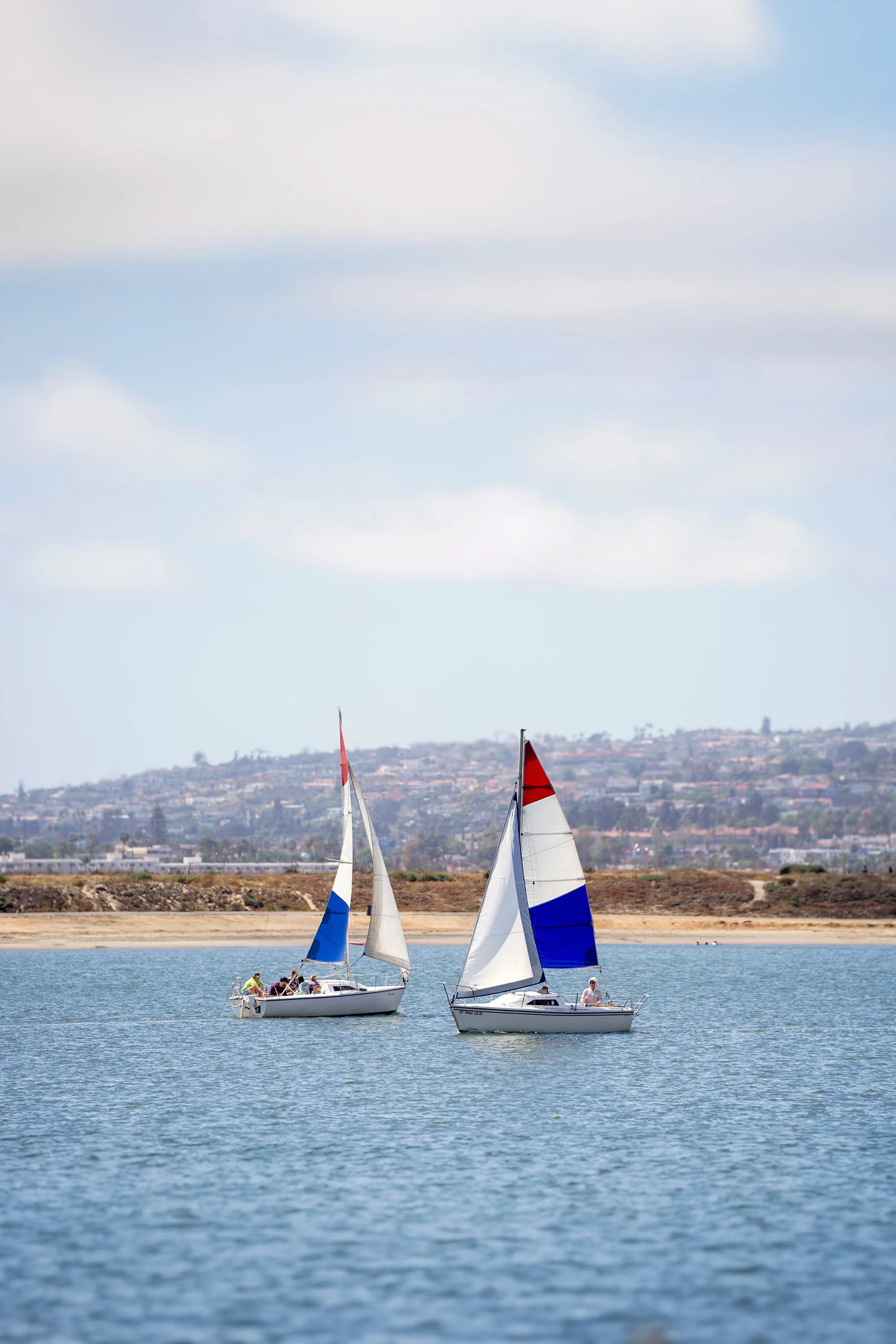 A two-person sailboat with a red, white, and blue sail glides across the calm water of Mission Bay in San Diego. In the background, a low-lying sandy shoreline sits before a hillside dotted with coastal homes. The scene is captured under a bright, ov