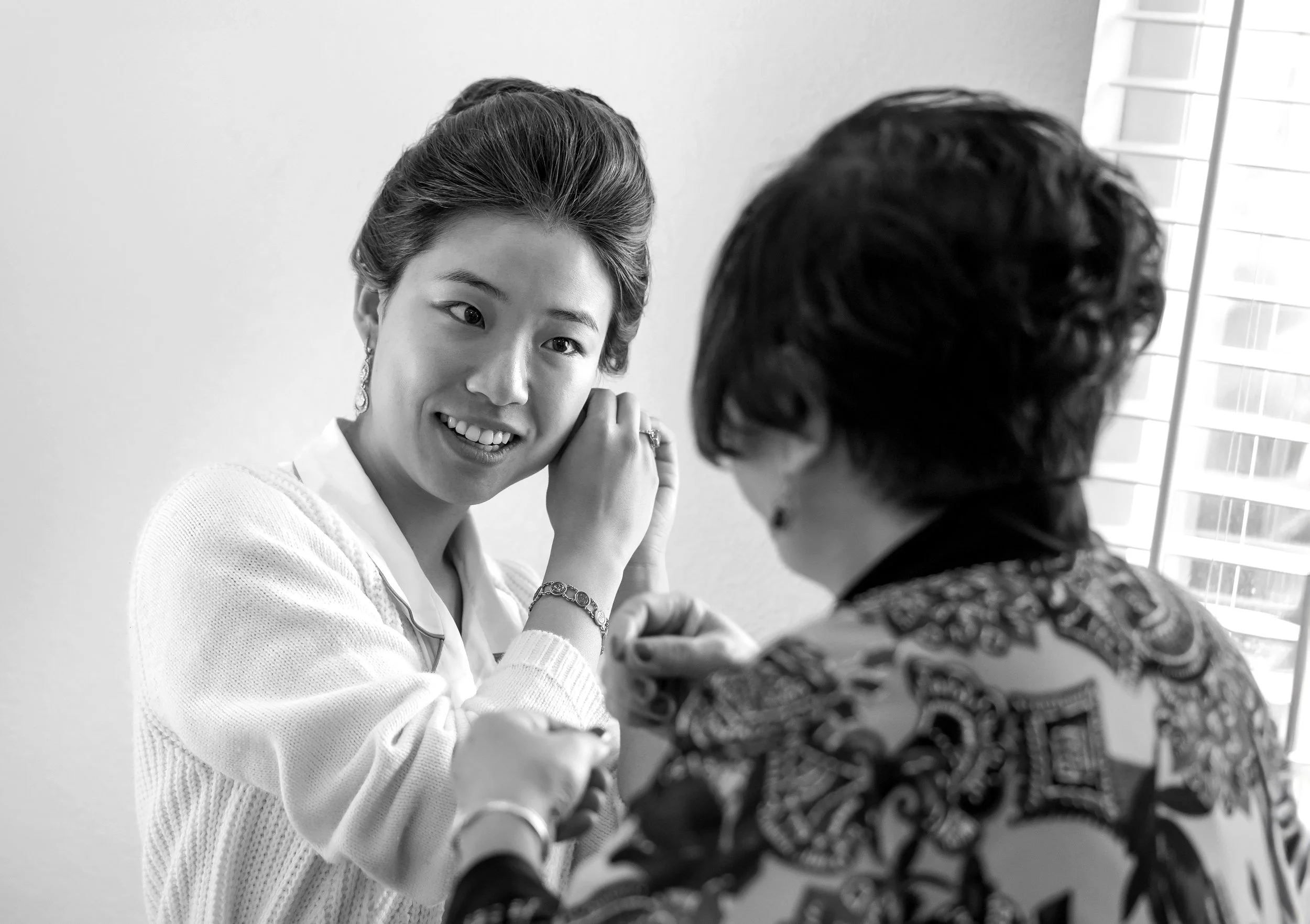 A black and white photo shows a bride with her dark hair in an updo smiling as an her mom, seen from behind, helps her put on an earring. The young woman is wearing a light-colored collared shirt and cardigan, a bracelet, and drop earrings. The older