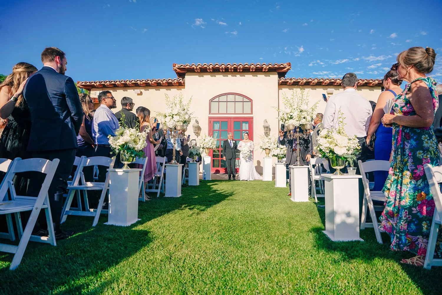 An outdoor photograph of a wedding ceremony on a sunny day in Maderas Golf Club. A bride in a white wedding dress and a groom in a dark suit stand at the altar area in front of a building with Spanish-style architecture and a red-tiled roof. Guests s