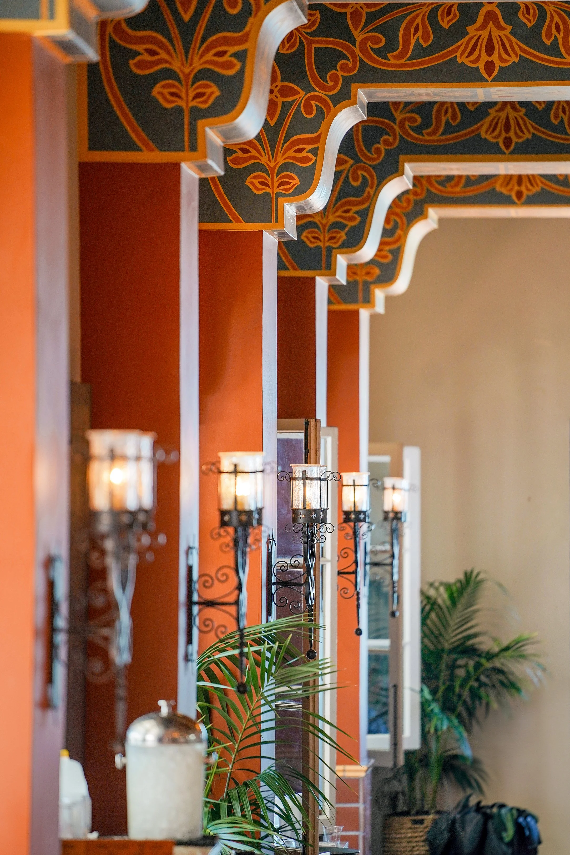 A hallway or entryway featuring Spanish Colonial-style architectural details, including painted ceiling beams with decorative scrollwork, orange-terracotta walls, black wrought-iron wall sconces, potted palm plants in baskets, and a glass beverage di