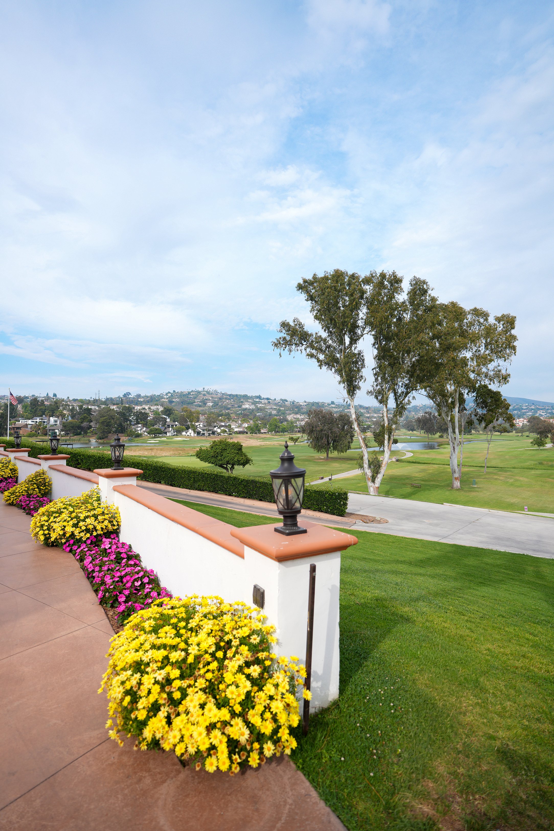 A scenic view from a patio overlooking a green golf course and rolling hills with houses, featuring vibrant yellow and pink flower beds and decorative black lanterns.