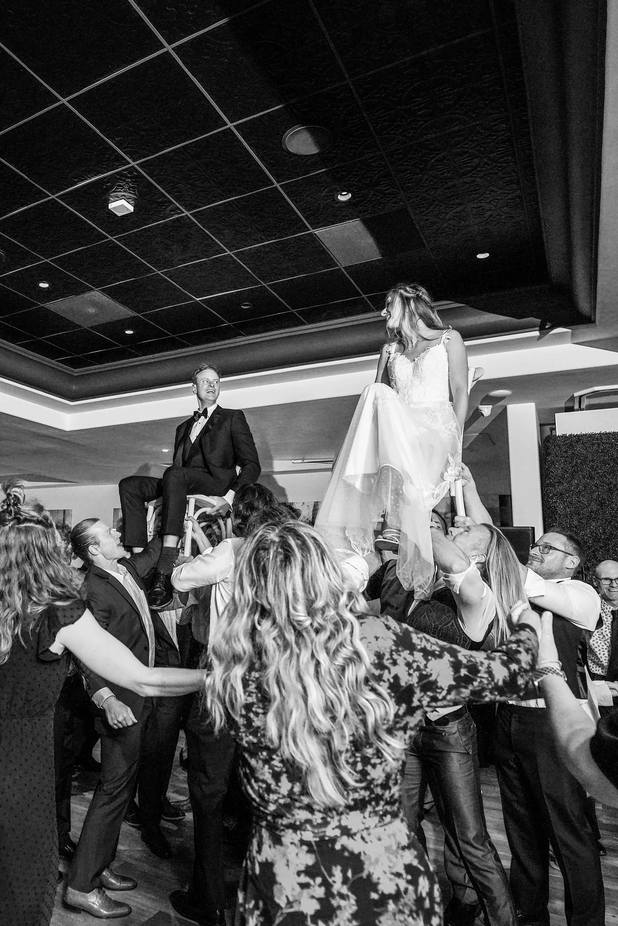 A black and white photo of a bride in a white dress and a groom in a tuxedo being lifted into the air on chairs by a crowd of wedding guests during a traditional dance.


