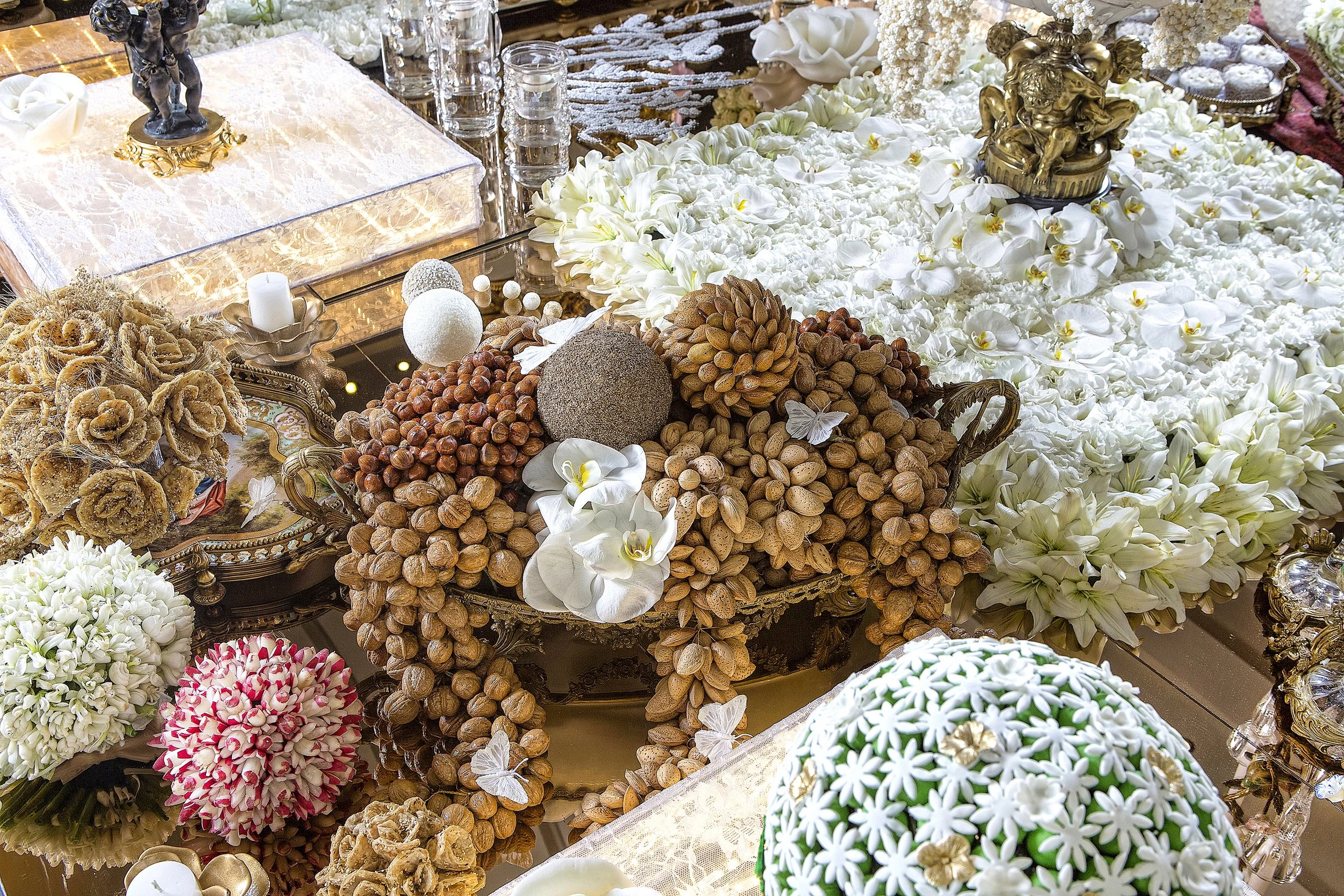 An ornate Persian Sofreh Aghd wedding ceremony table featuring symbolic items including nuts, sweets, an ornate gold clock, miniature bride and groom figures, and extensive white floral arrangements with gold accents.