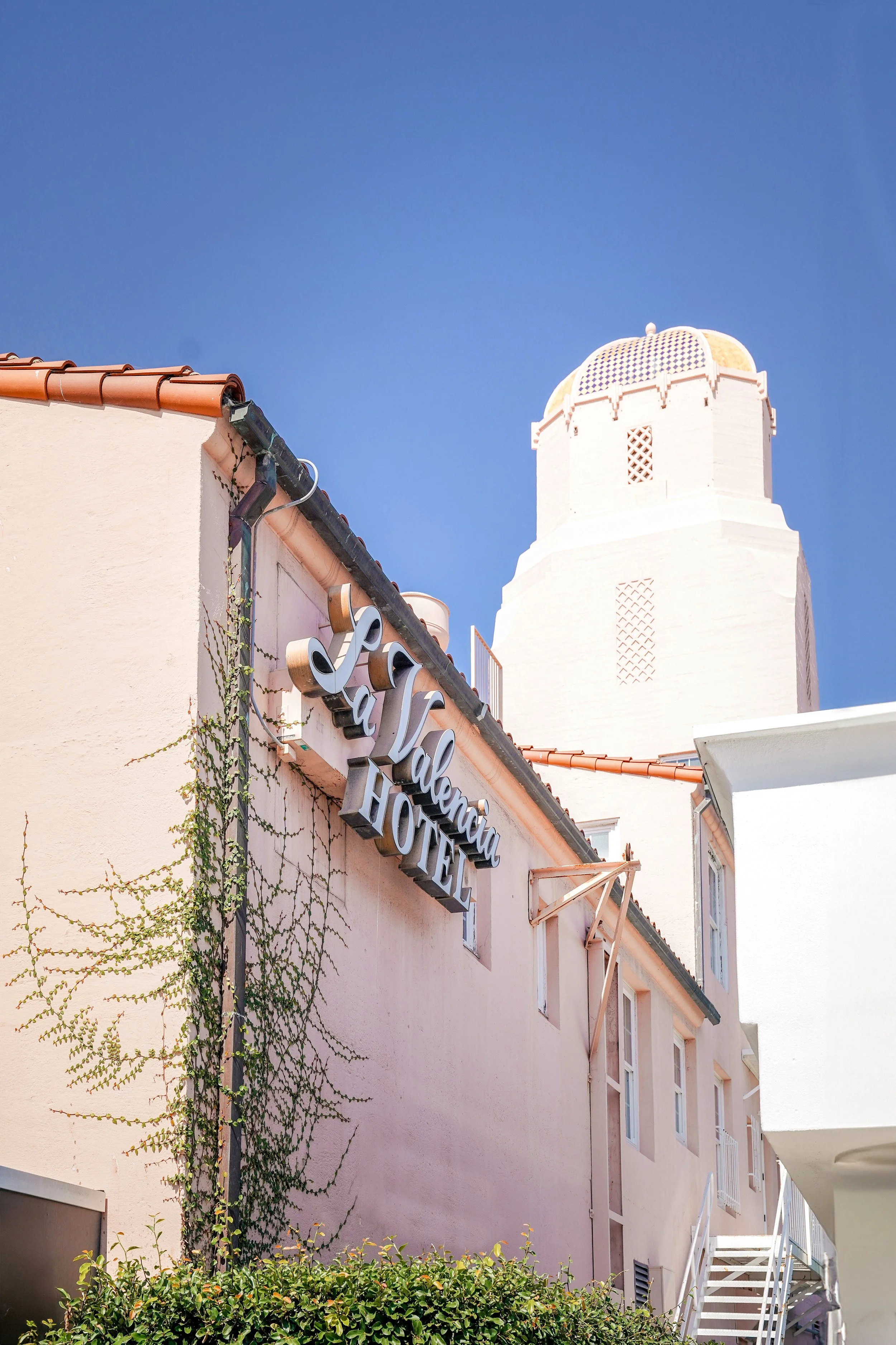 A close-up view of the pink-stucco building facade of the La Valencia Hotel, featuring an ornate "La Valencia Hotel" sign, a white architectural tower in the background, climbing ivy, and a clear blue sky.
