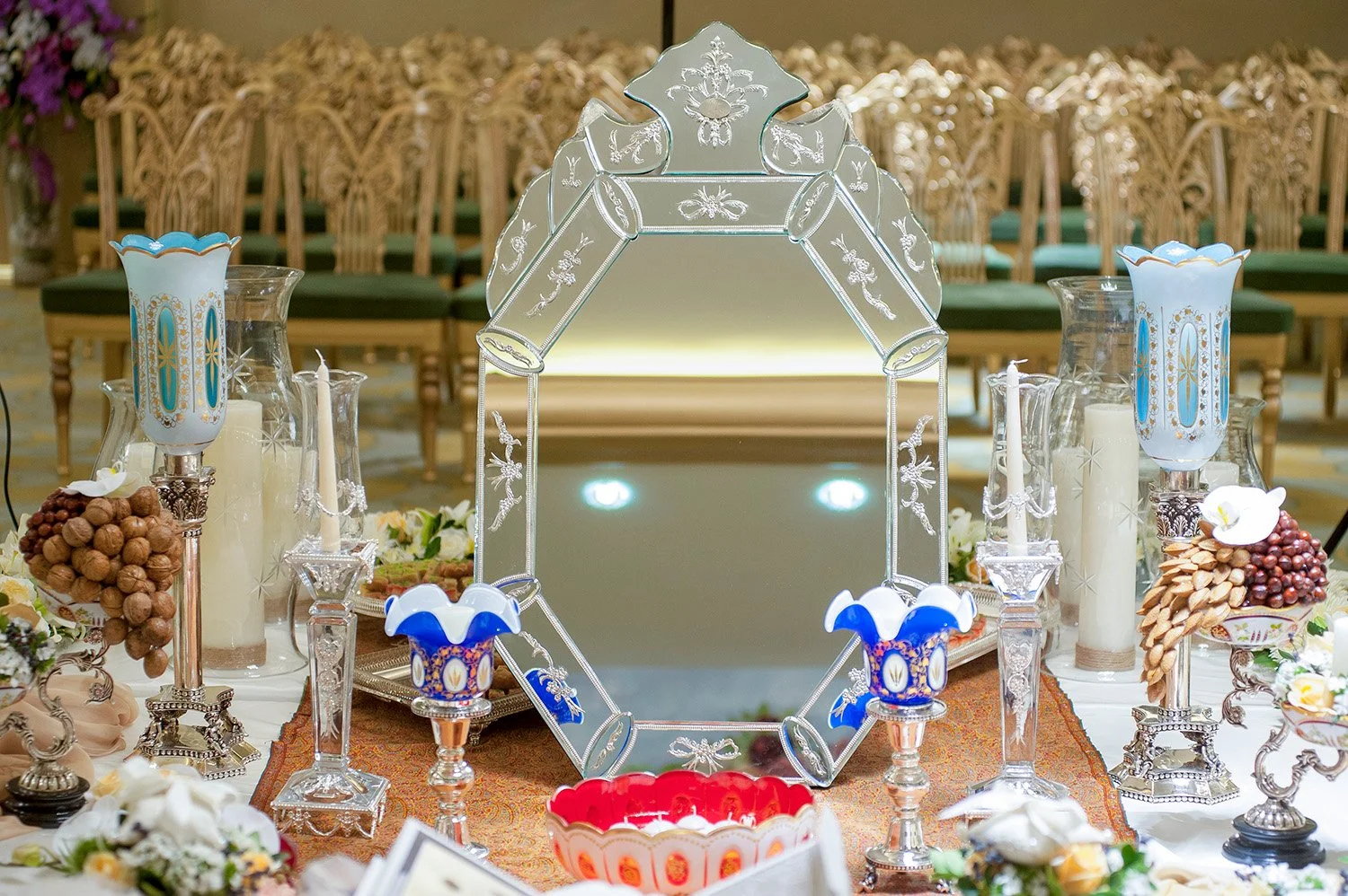 An elaborate, formal indoor table setting for a Persian wedding ceremony (Sofreh Aghd) featuring a large ornate mirror, silver candelabras with blue glass, nuts, candles, and white flowers.