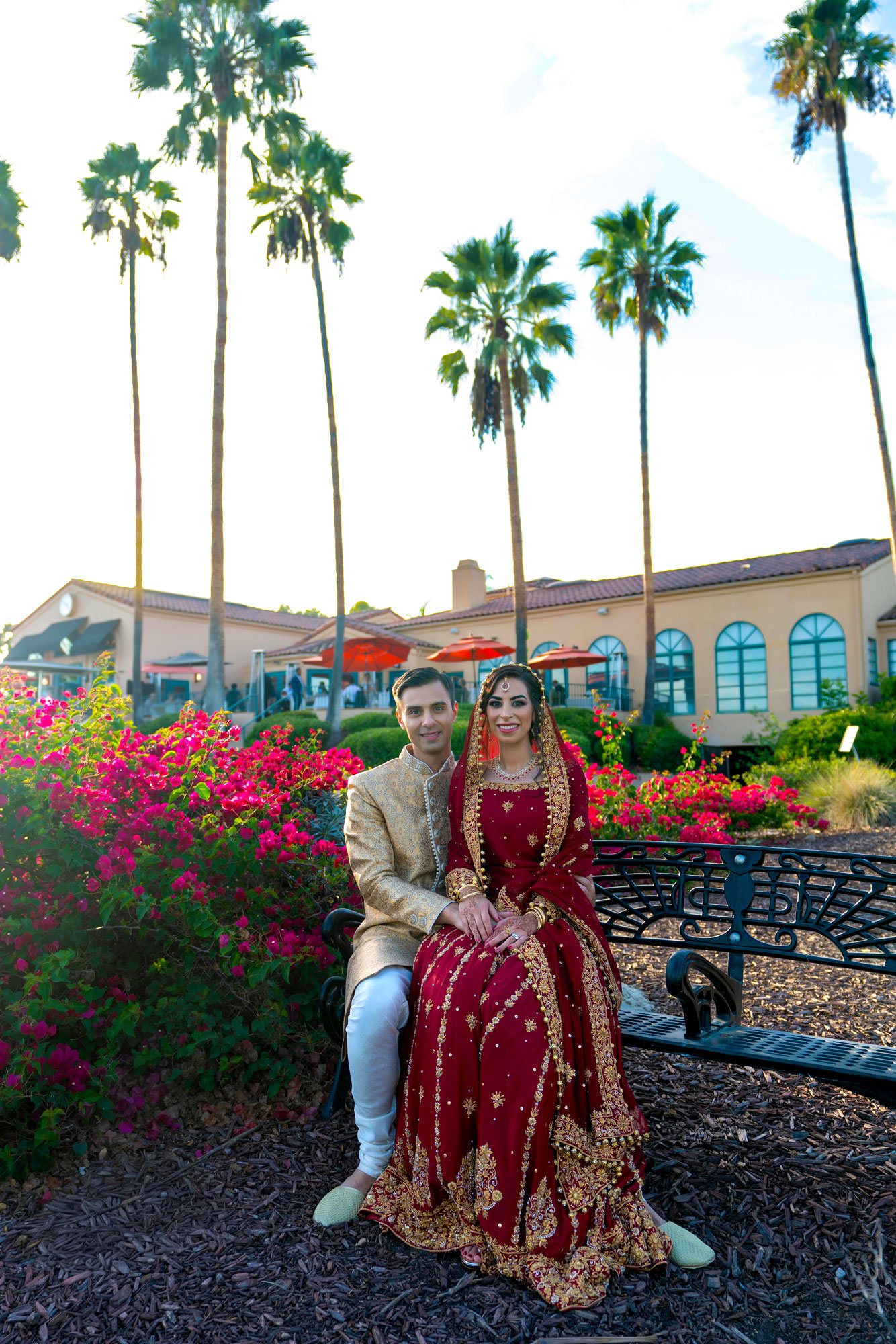 A photograph of a newly married couple in traditional Indian wedding attire, seated on a black metal bench outdoors. The groom wears a gold sherwani and white pants, and the bride wears an ornate, deep red lehenga with gold embroidery and a veil. The