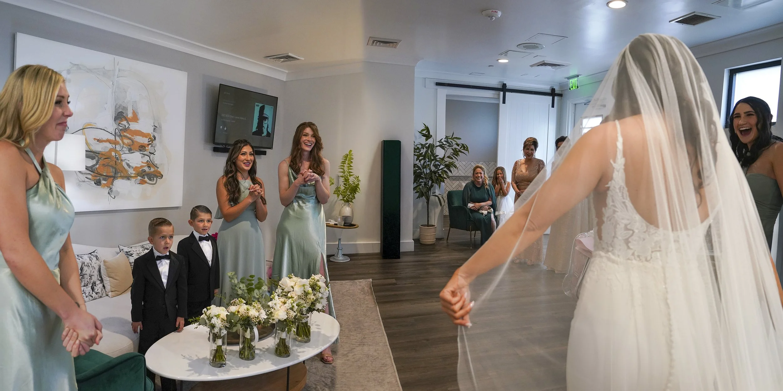 In a bright, modern room, a bride in a white, lace-bodice wedding gown and a long, sheer veil stands with her back to the camera, showcasing her dress to a group of friends and family. To her left, two young boys in small black tuxedos and bowties lo