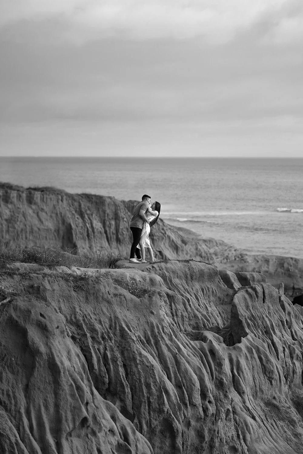 A black and white photograph of a couple embracing and kissing on a rugged, rocky cliff overlooking the ocean. The man wears a light shirt and dark pants, and the woman wears a light-colored dress. The sky is overcast.