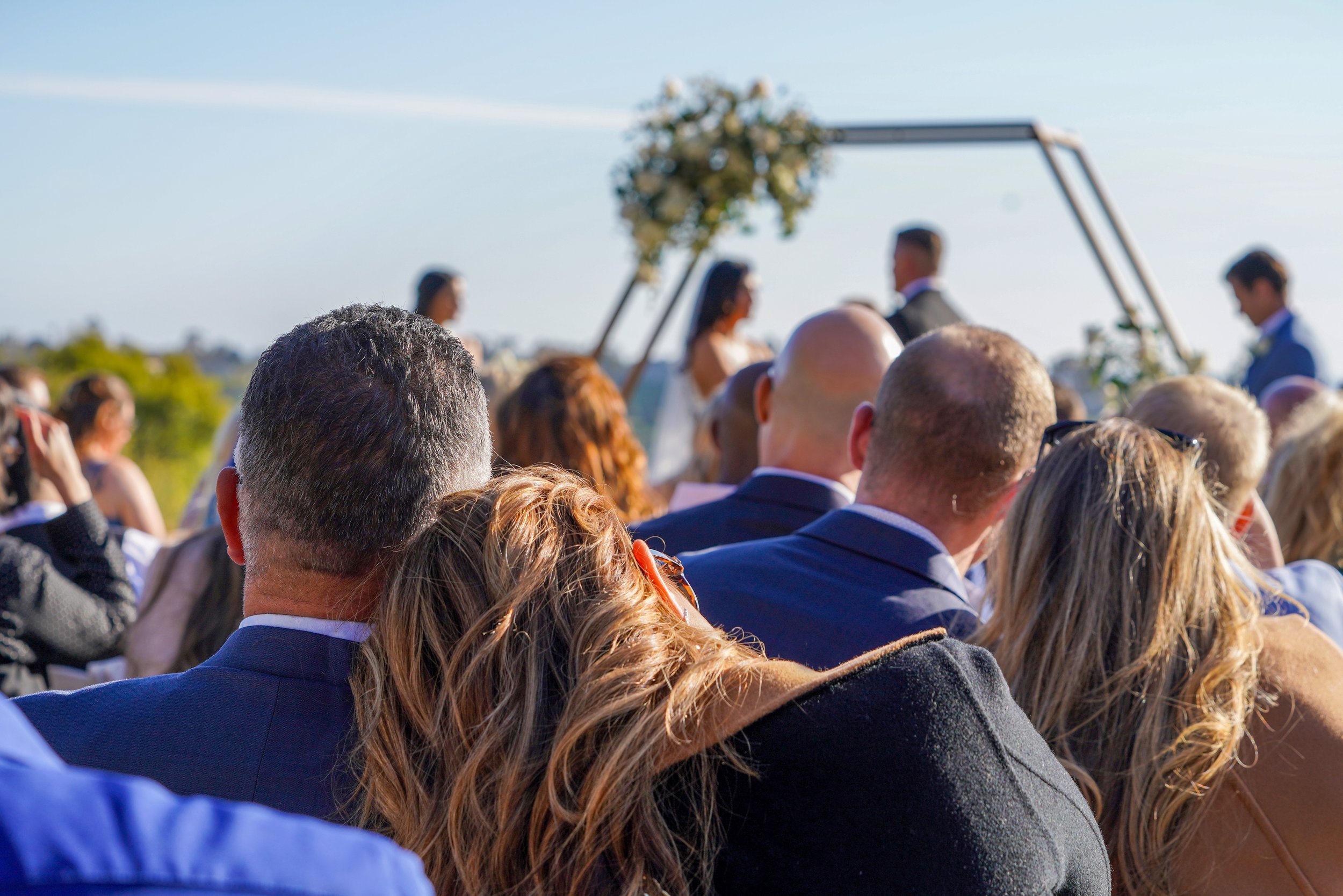 Over-the-shoulder view of wedding guests watching a bride and groom during an outdoor hilltop ceremony with a hexagon floral arch.