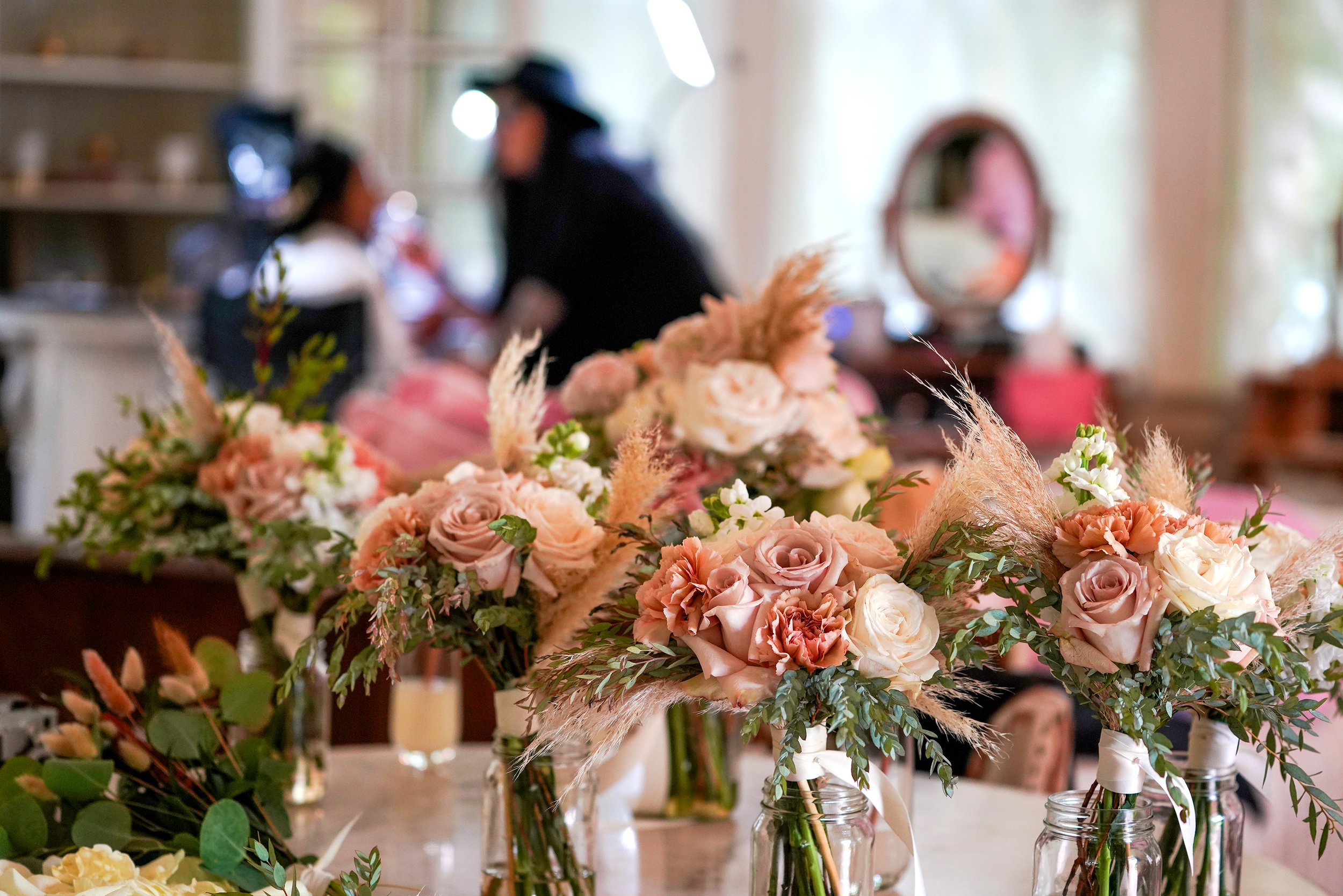 Several rustic-style wedding bouquets featuring dusty rose, peach, and white roses with pampas grass, displayed in glass jars on a table with a blurred background of a bridal suite.