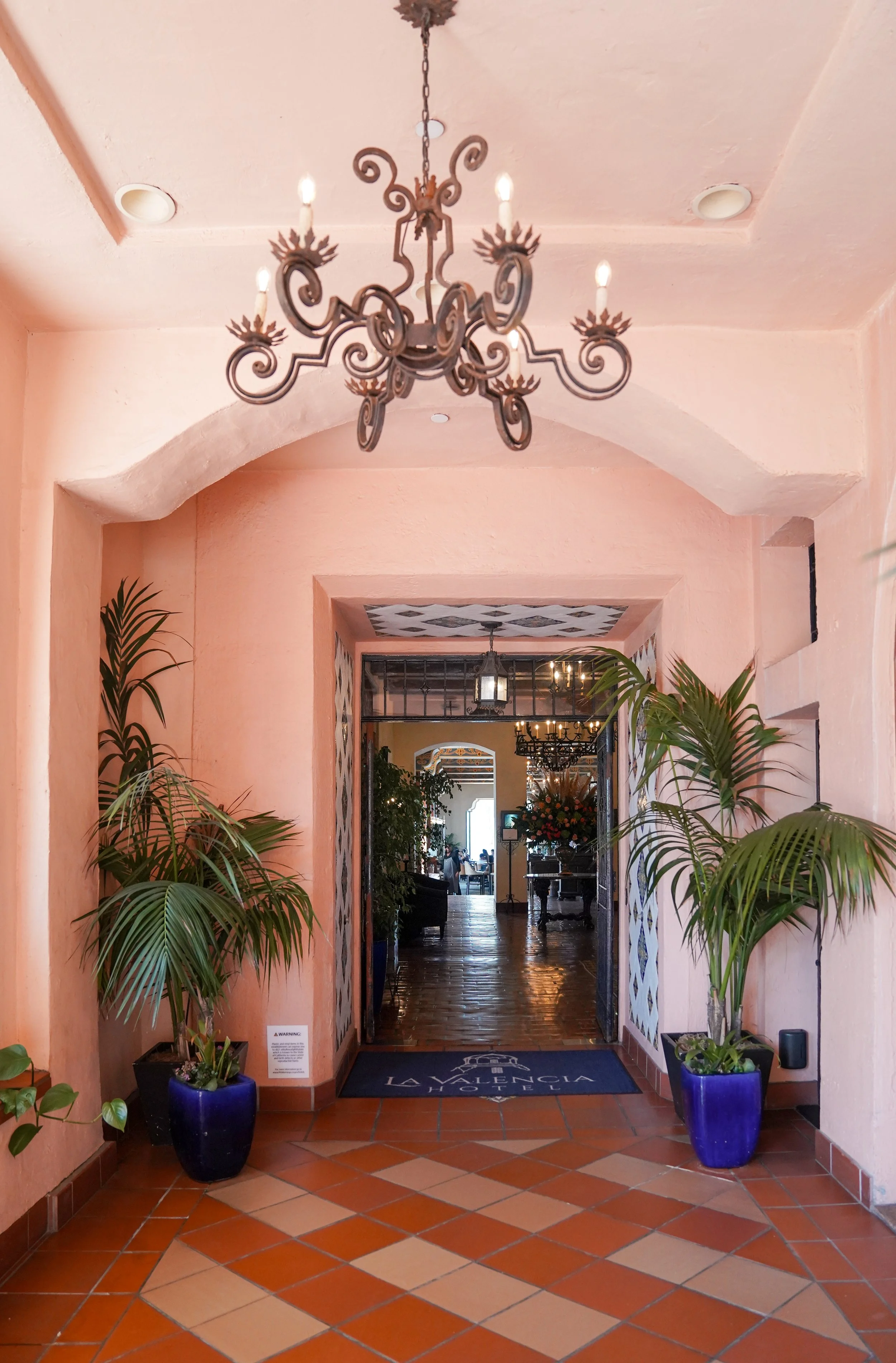 The entrance to the La Valencia Hotel featuring an arched doorway framed by pink stucco walls and blue potted palm plants, terracotta tile flooring, an ornate wrought iron chandelier, and a view into the hotel lobby.
