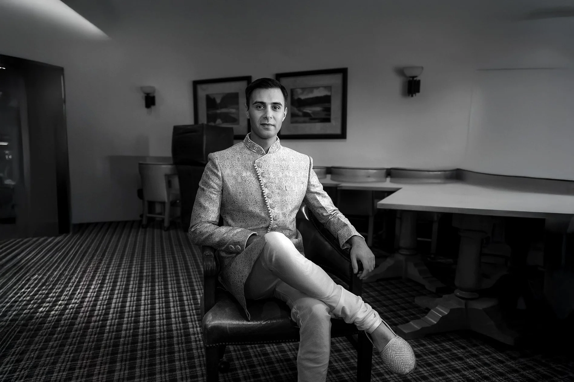 A black and white photograph of a groom in traditional Indian formal wear, seated in an indoor setting. He is wearing a patterned sherwani jacket, light trousers, and decorated slip-on shoes. He sits with his legs crossed in a dark leather chair, loo