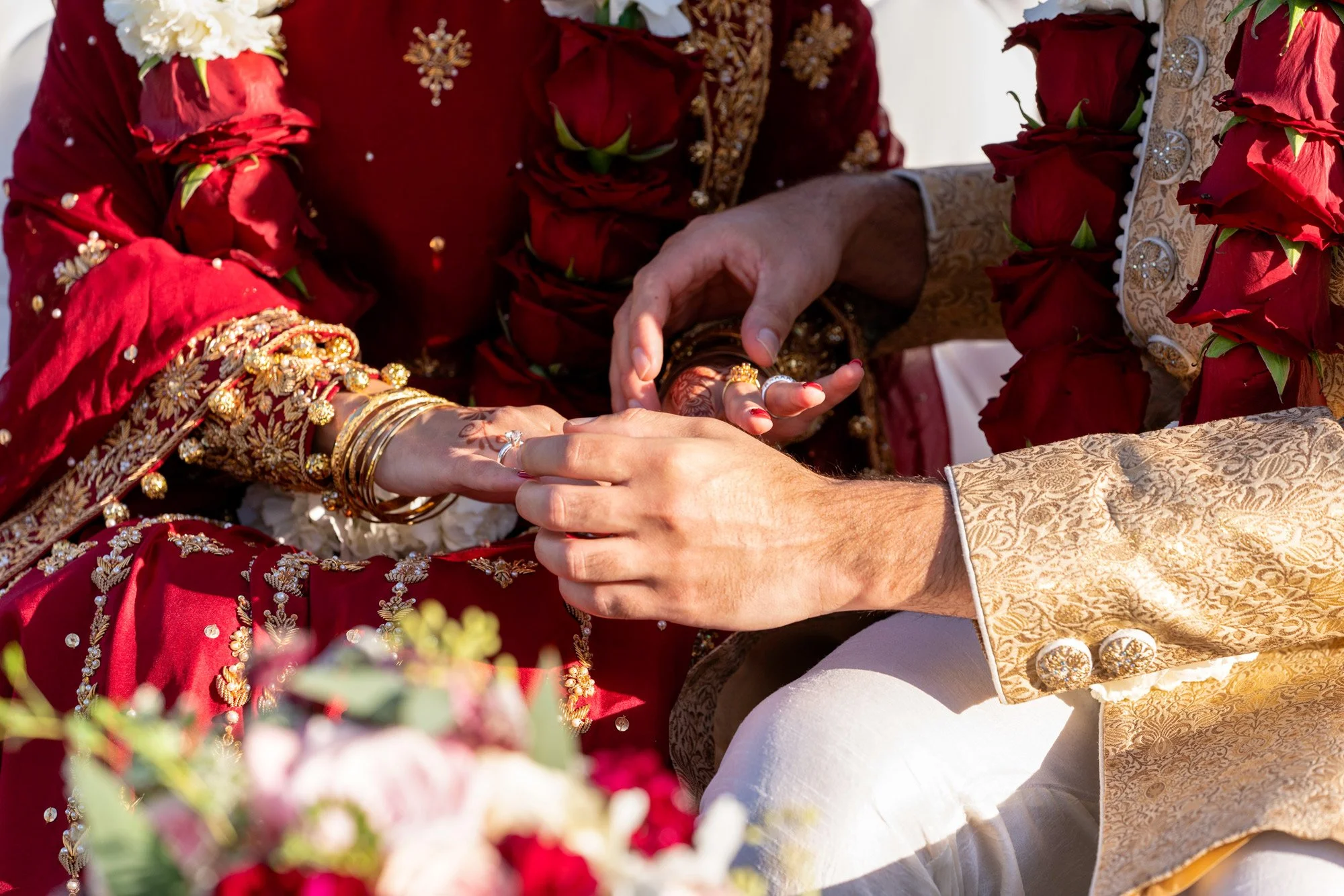 A close-up photograph capturing a groom placing a wedding ring on his bride's finger during a ceremony. The bride wears a traditional deep red and gold outfit with several gold bangles on her wrist, and the groom wears a gold brocade sherwani. Red ro