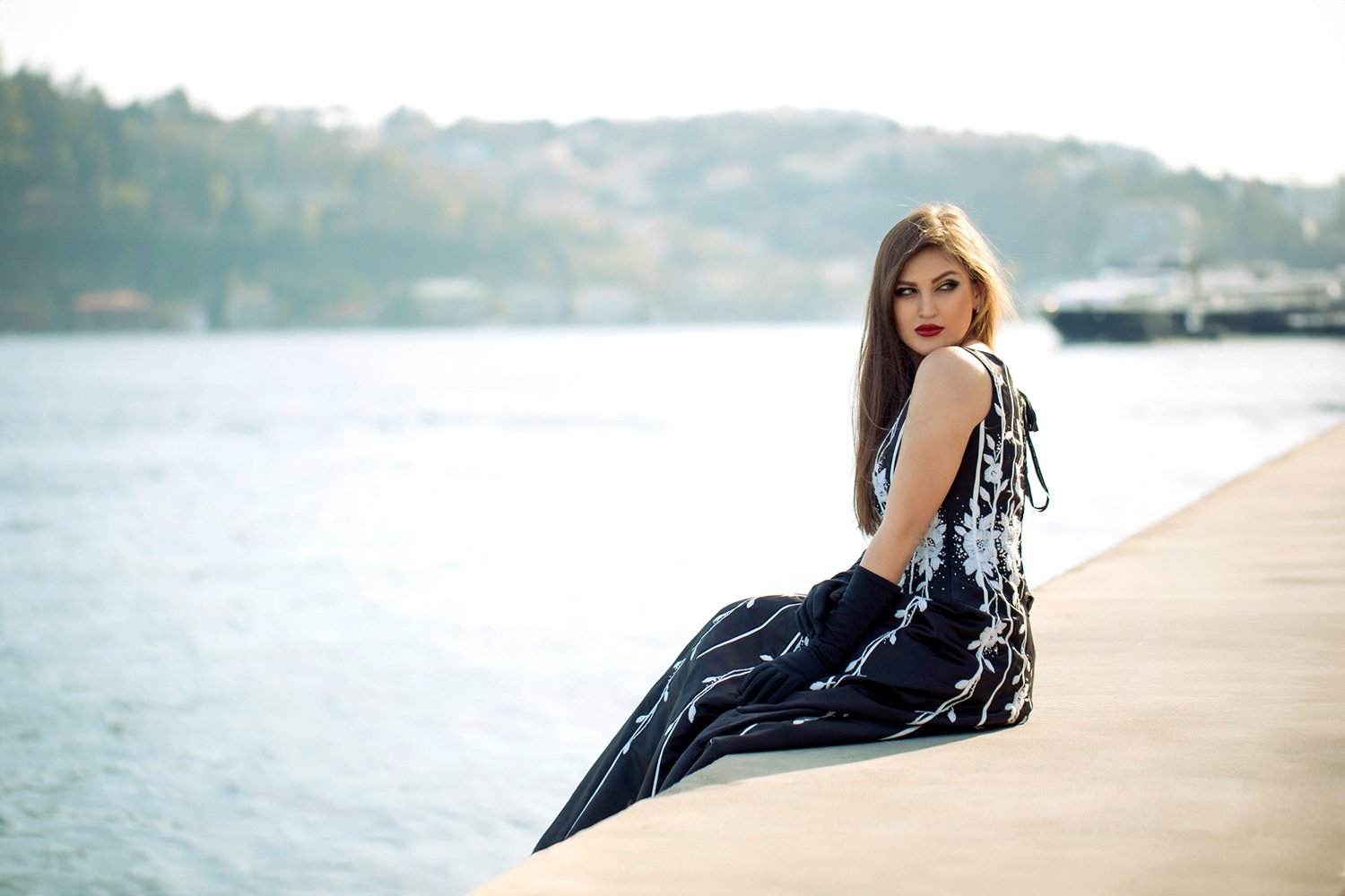 A young woman in a black and white floral dress and black gloves is sitting on a concrete dock by the water, with boats and a distant shoreline in the background.