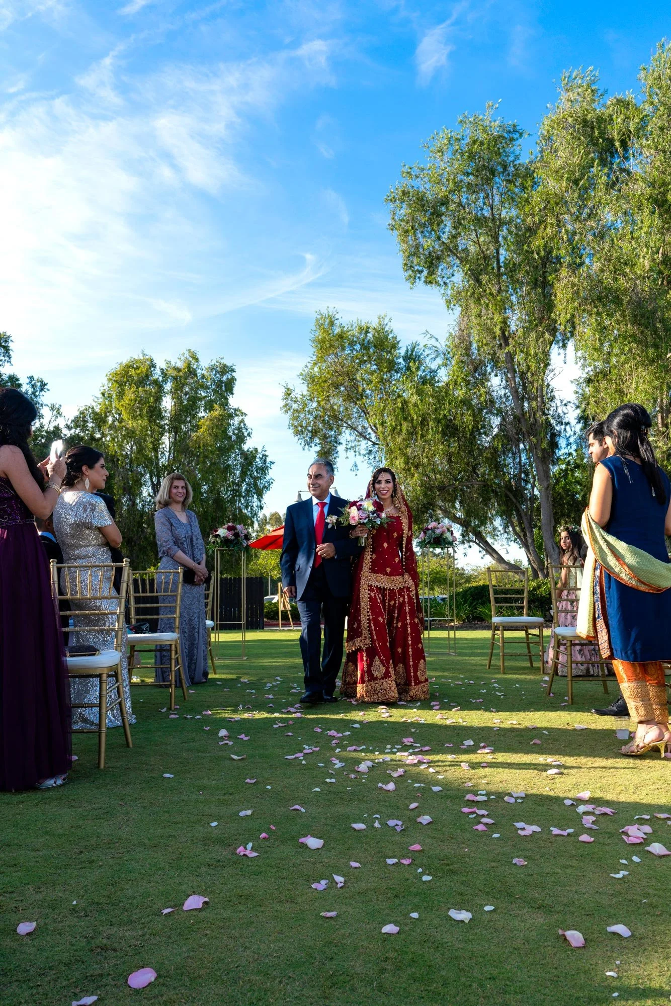 An outdoor photograph of a wedding procession on a sunny day. An older man in a navy suit and red tie escorts a bride in an ornate, traditional red and gold lehenga down a grassy aisle scattered with pink and white rose petals. Guests seated on gold 
