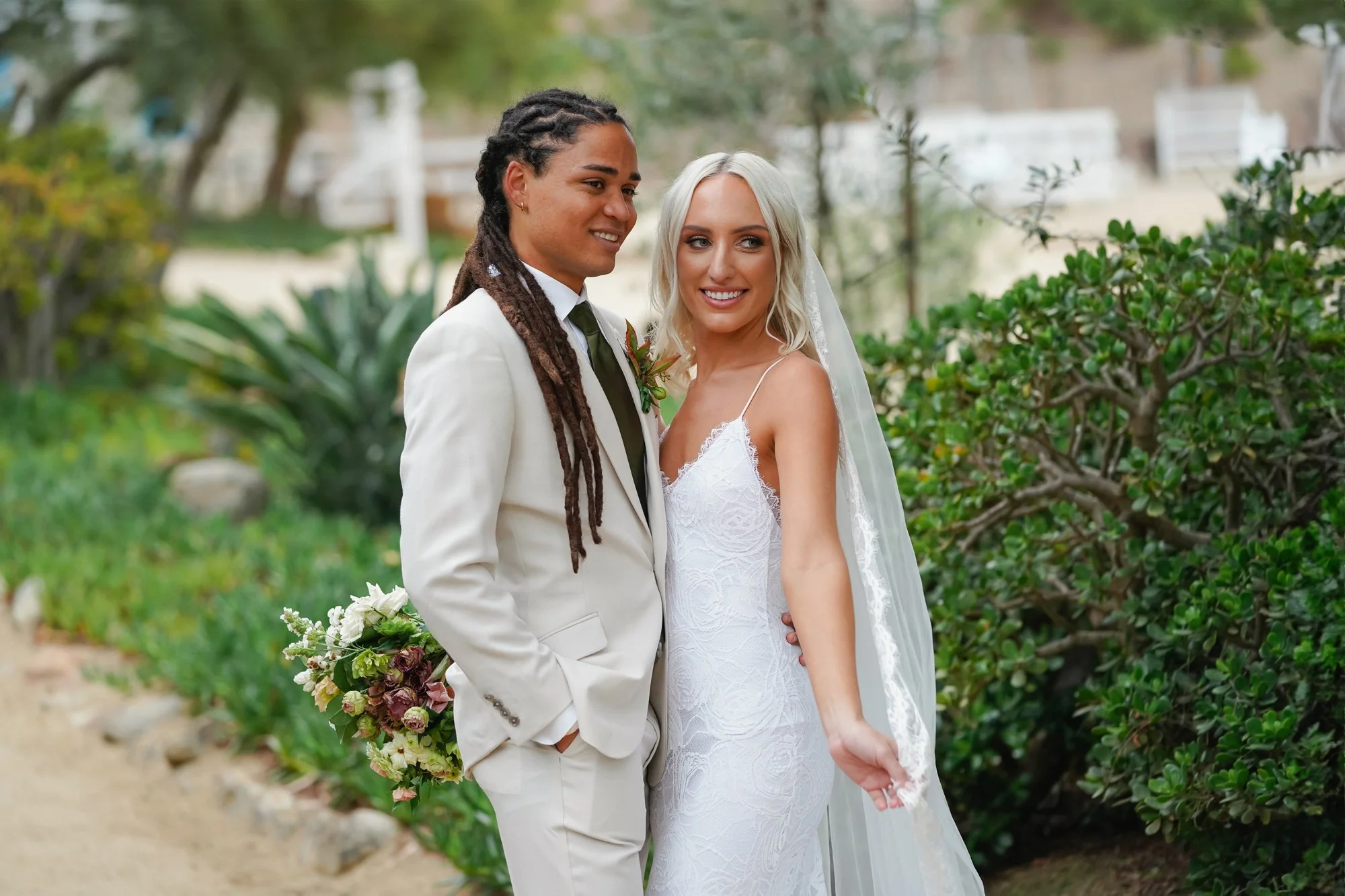 A photo of a newly married couple posing for a portrait, with the groom in a cream-colored suit looking towards the bride who is in a white lace slip dress and veil and smiling happily at the camera.


