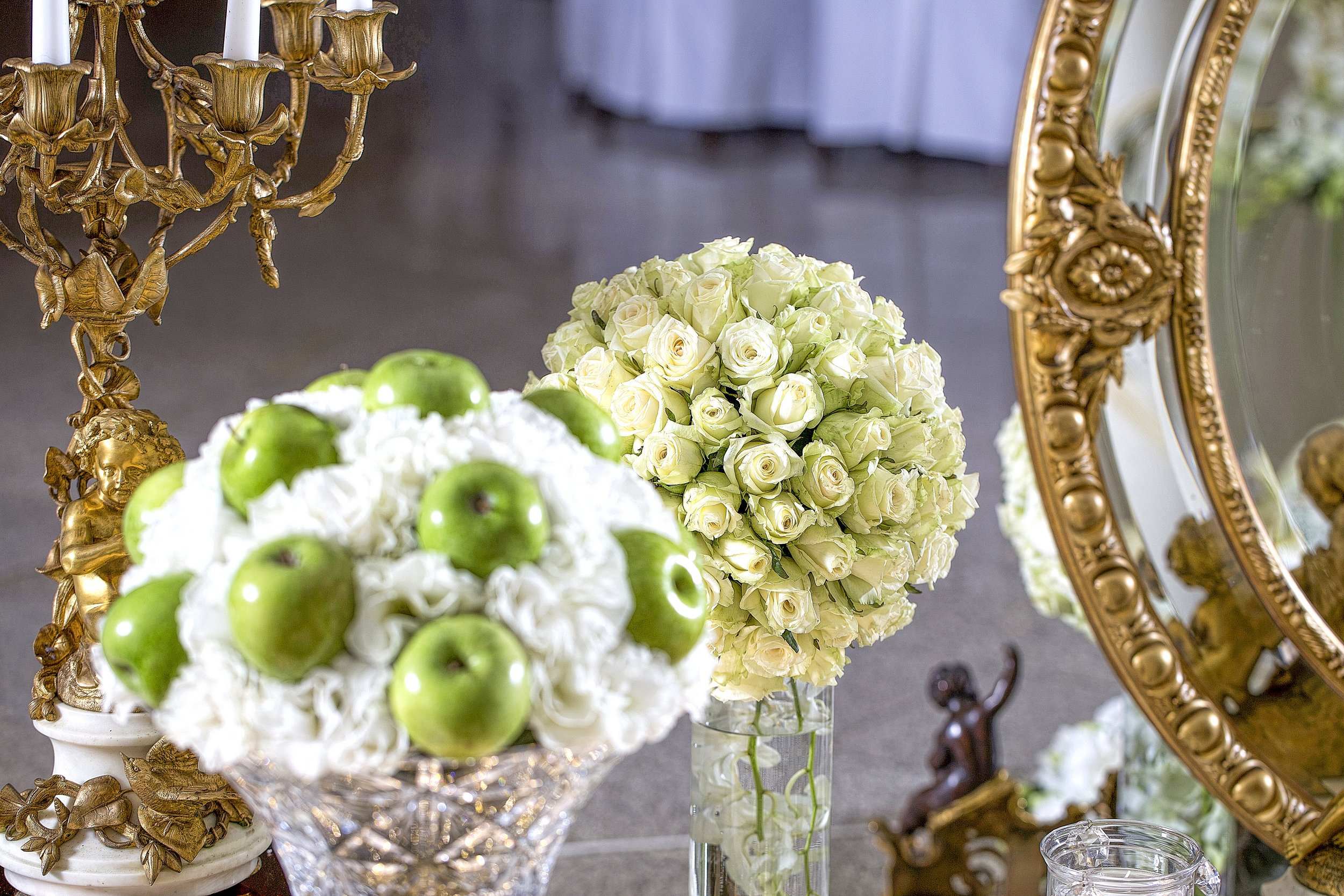 A close-up view of two floral arrangements, one featuring green apples mixed with white flowers and another with a sphere of cream roses, positioned next to an ornate gold candelabra and a large gold-framed mirror.
