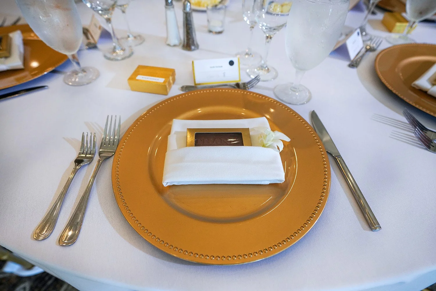 A formal dining table place setting featuring a gold charger plate, a white napkin holding a gold-wrapped favor, silver forks and a knife, clear water and wine glasses, and a place card on a white tablecloth.
