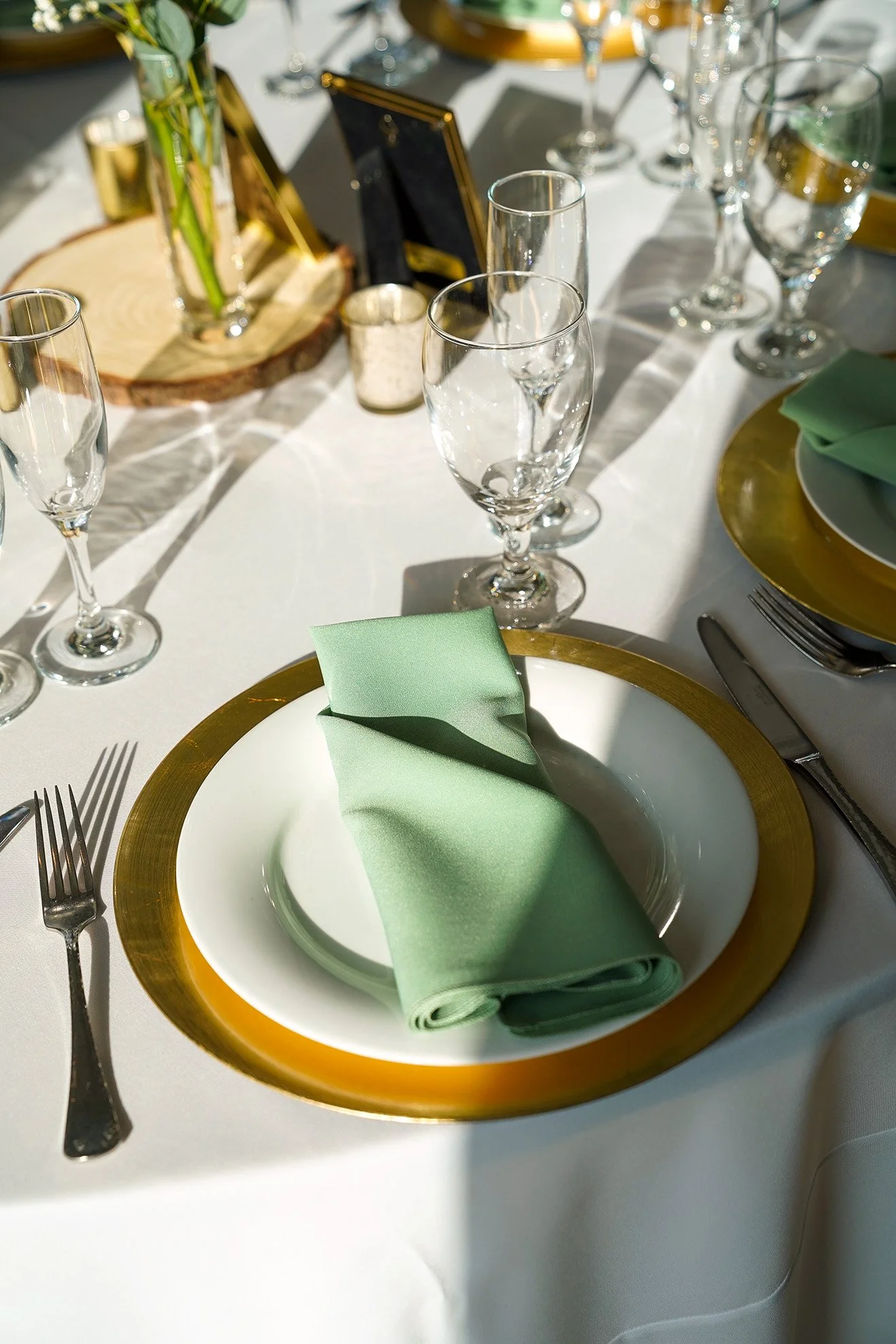 A formal dining table place setting featuring a gold charger plate, a white plate, a green cloth napkin, silver cutlery, wine glasses, and a wooden placemat in the background.