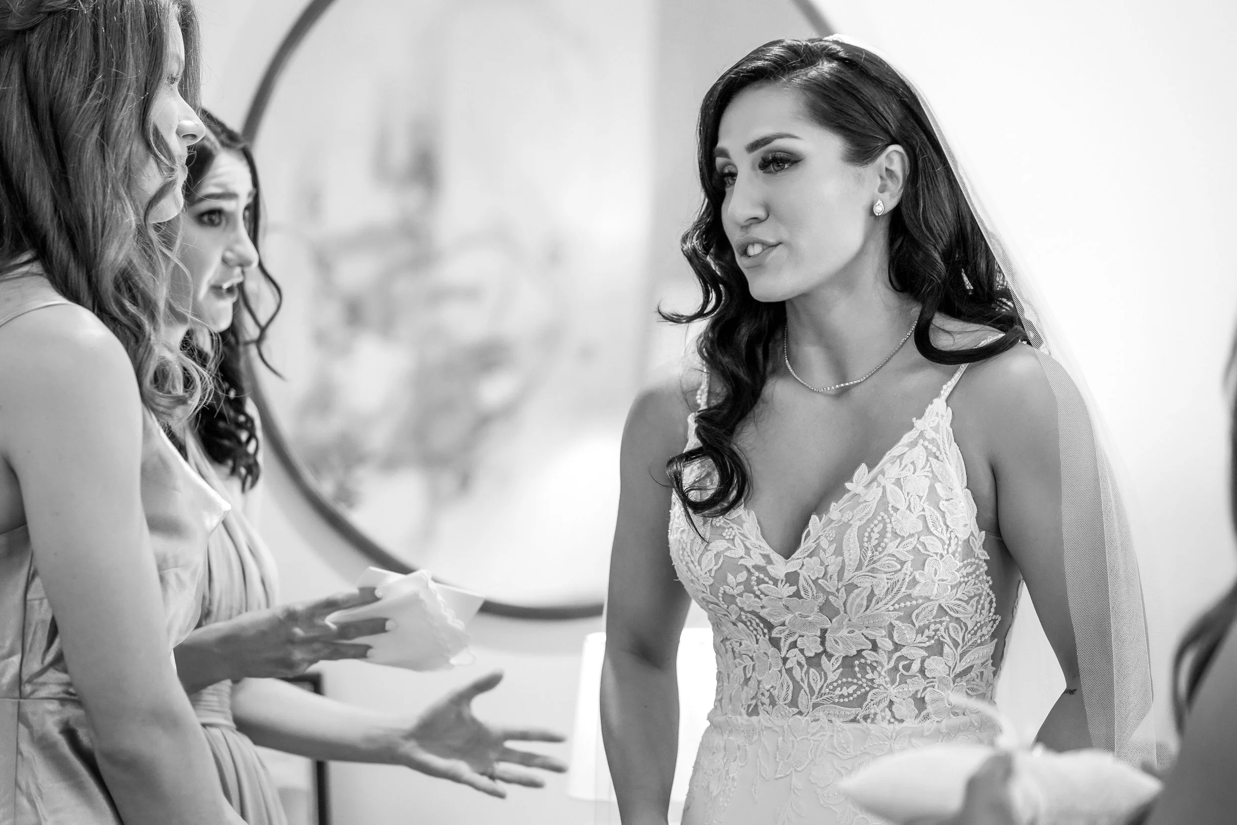 A joyful bride with long wavy hair and a delicate veil shares an emotional moment with her bridal party while getting ready for her wedding.